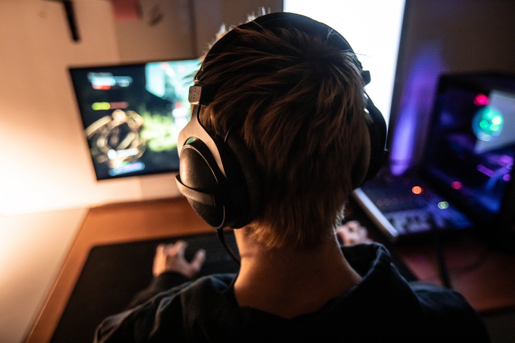 A person with headphones is seen from behind while playing a computer game, with a screen and gaming equipment in view. Names unknown