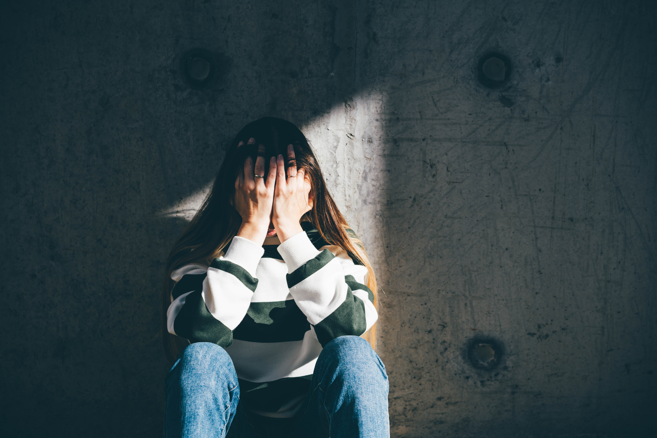 A person sits against a wall with their hands covering their face, appearing distressed. The person is wearing a striped long-sleeve shirt and jeans