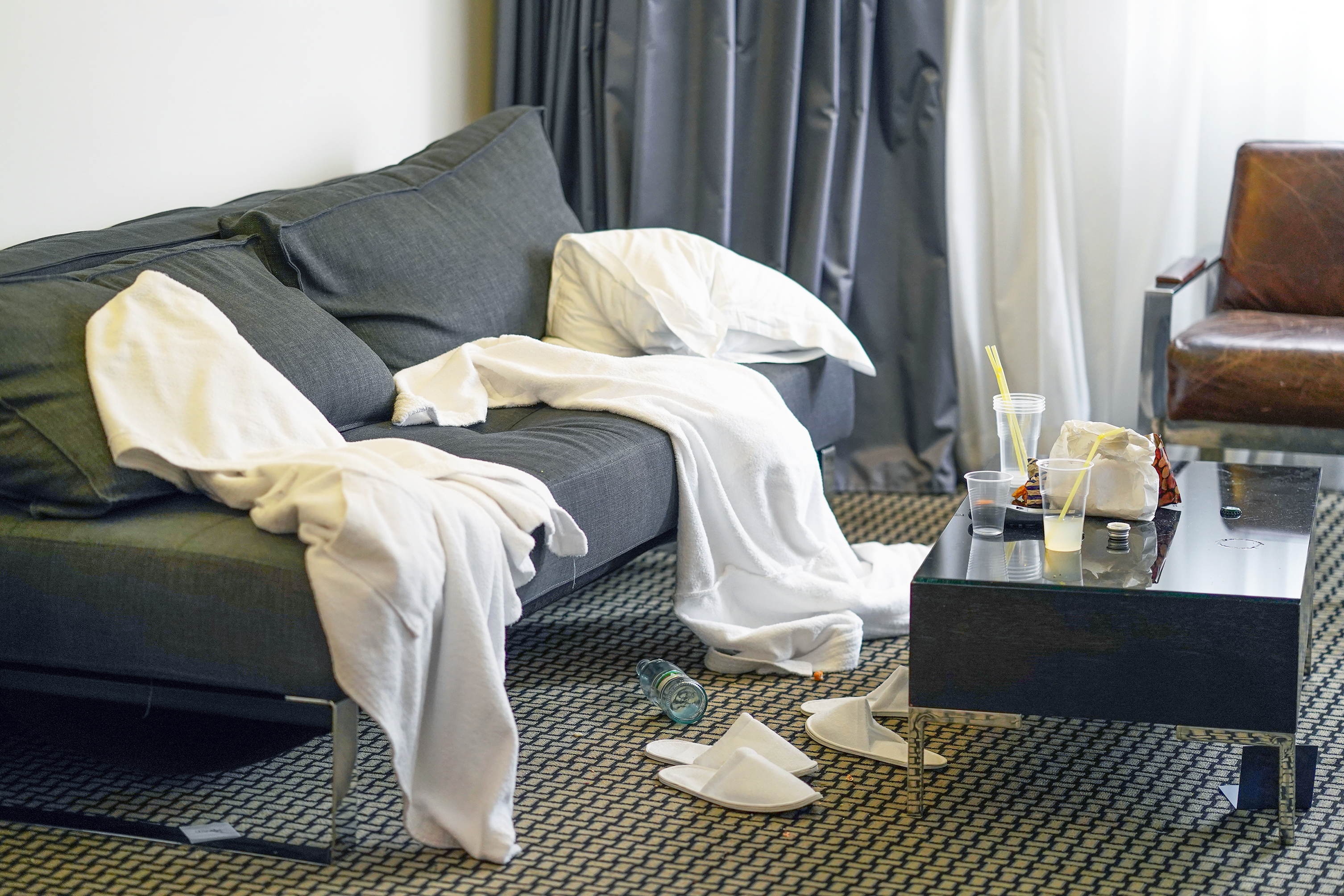 A messy hotel room with a couch covered in white towels and robes, a glass table with drinks and food items, and discarded paper items on the floor. No people present
