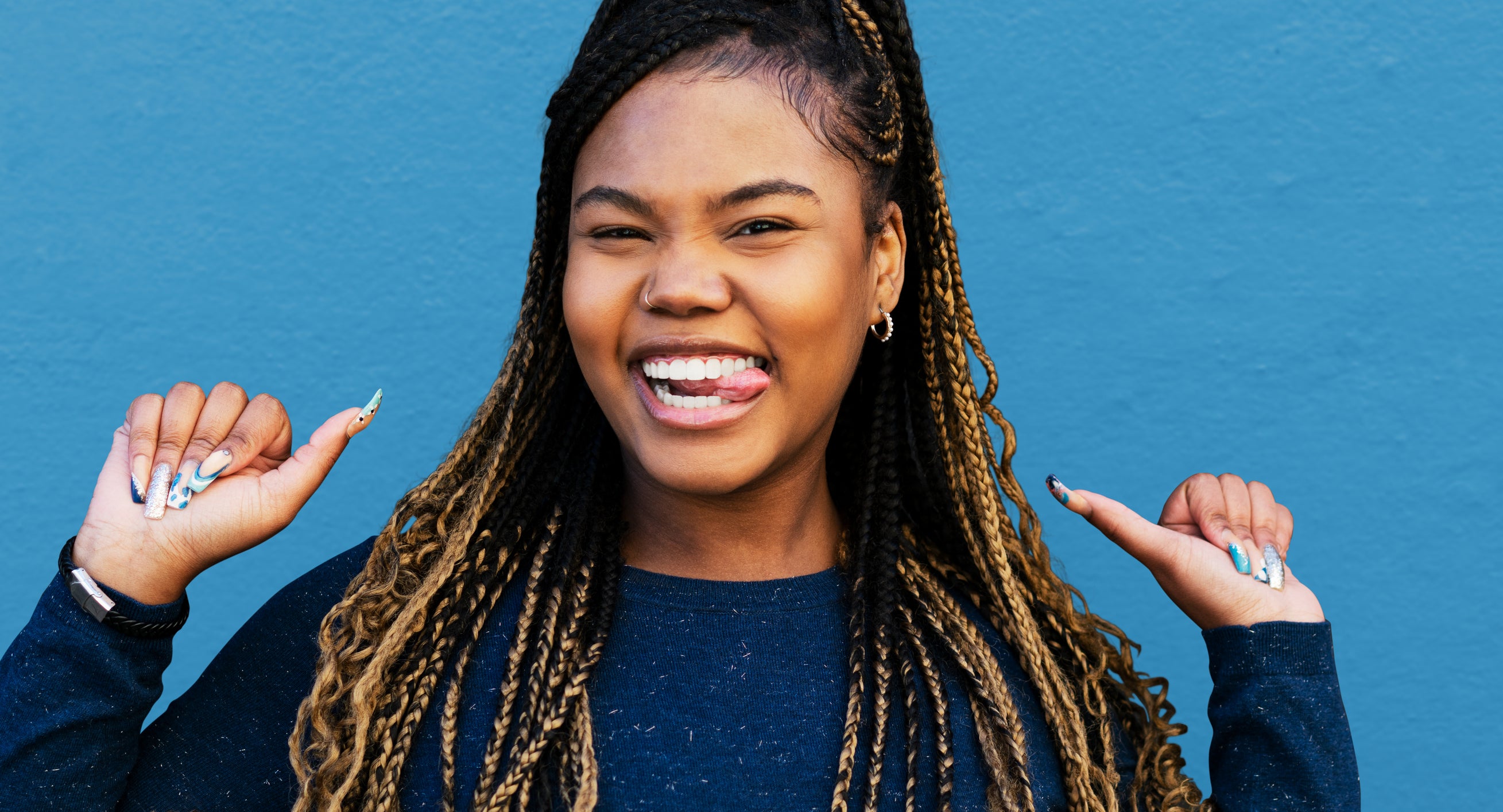 A woman with long braids, wearing a casual top, smiles brightly while pointing both thumbs toward herself in front of a plain background