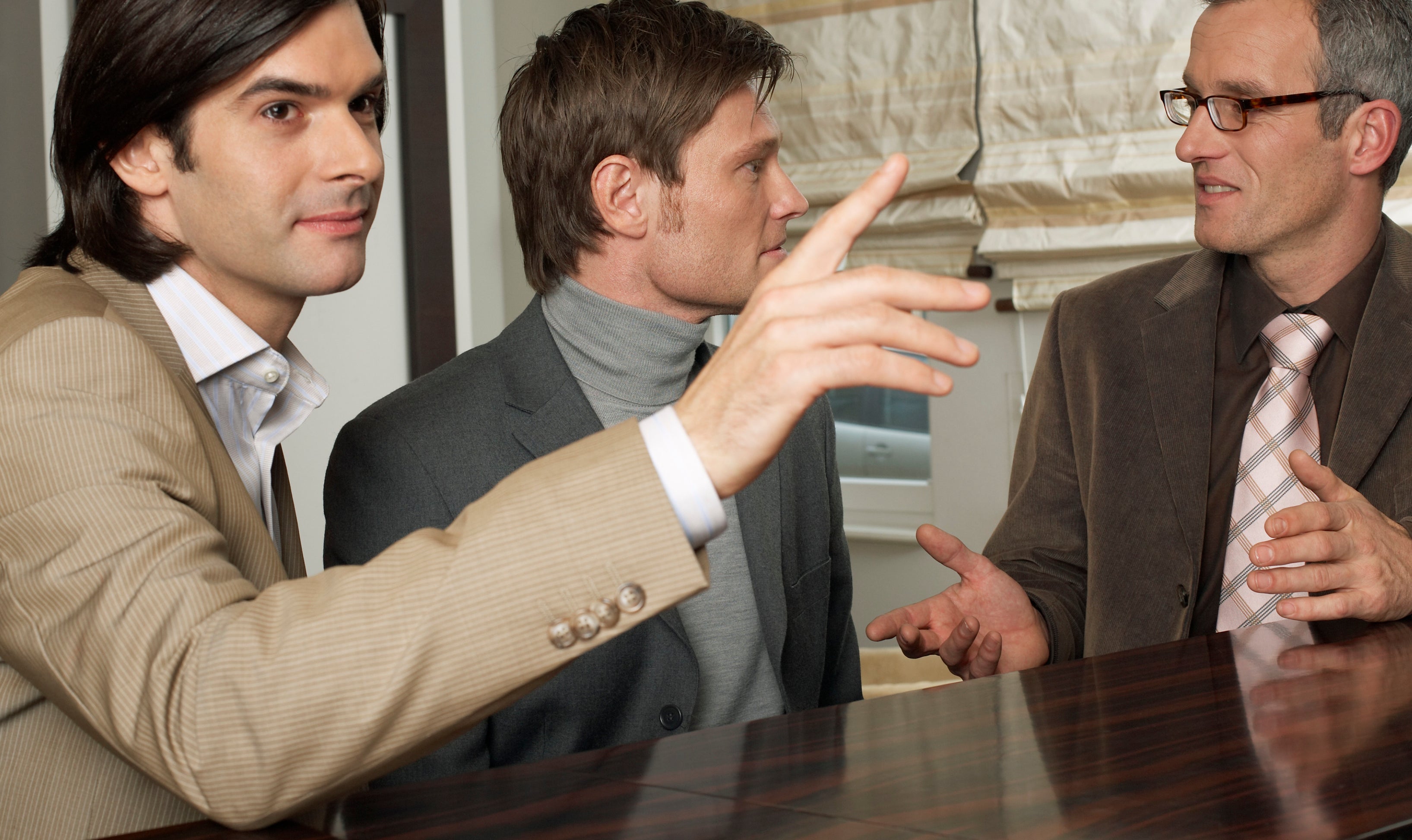 Three men, dressed in suits, engage in conversation at a bar counter