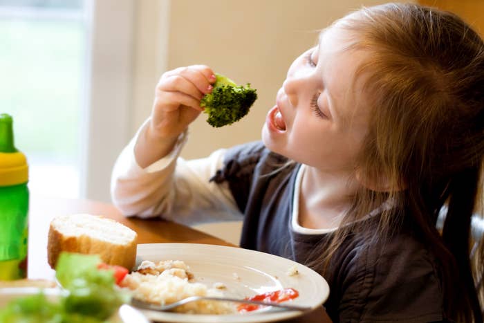 A young girl eats broccoli at a dining table with a plate of food, including bread and rice. A green sippy cup is next to her