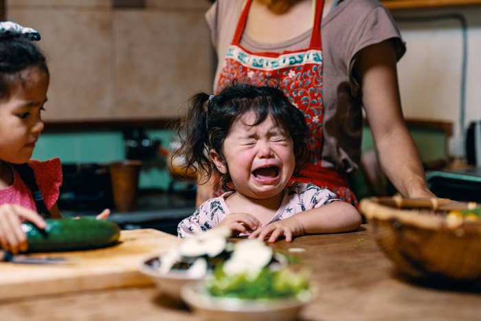 A young child is crying at a kitchen counter, while an adult in an apron stands behind them. Another child is chopping vegetables nearby