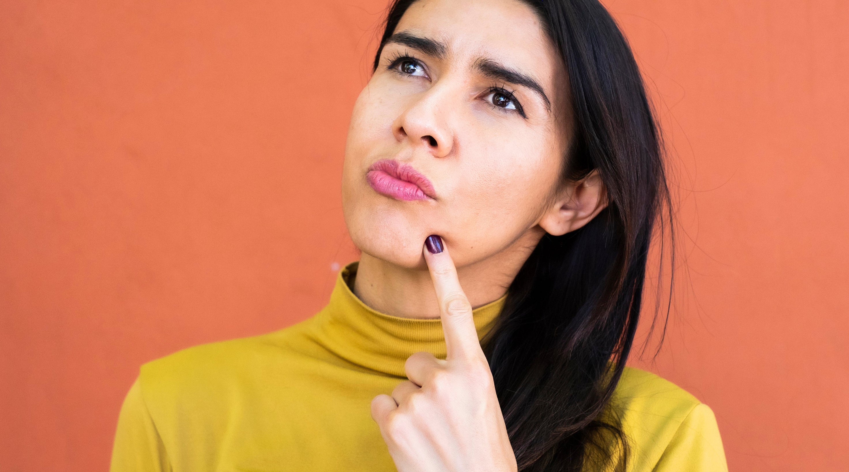 A woman in a yellow turtleneck looks thoughtful, resting her chin on one hand against a plain background