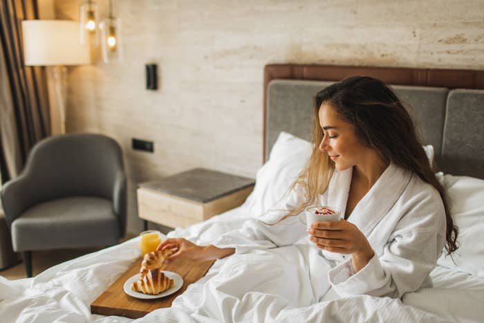 A woman in a white bathrobe sits on a hotel bed, holding a cup with yogurt and reaching for a plate of waffles next to a glass of orange juice