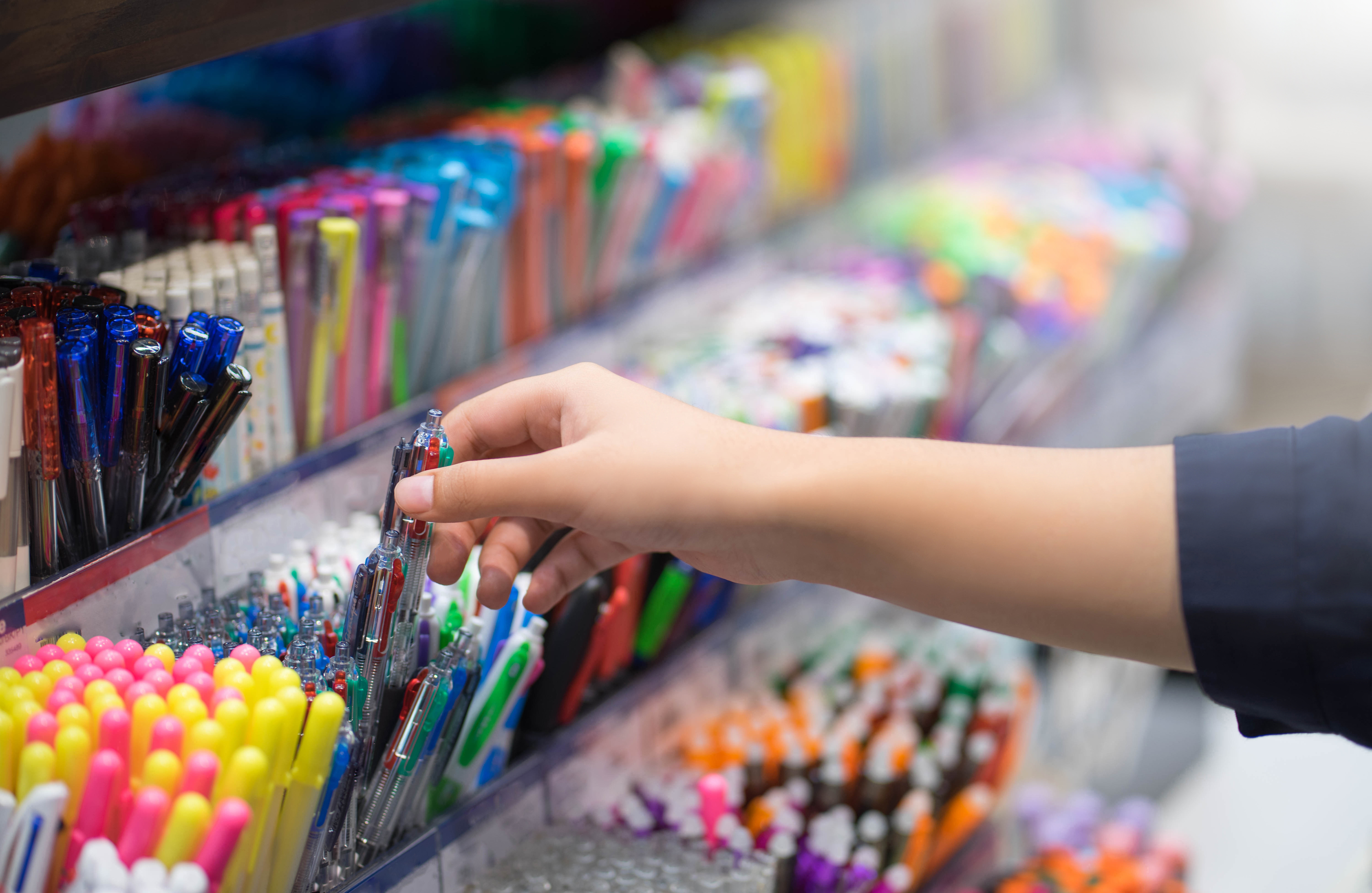 A hand reaches for a pen among a variety of writing instruments displayed on a store shelf