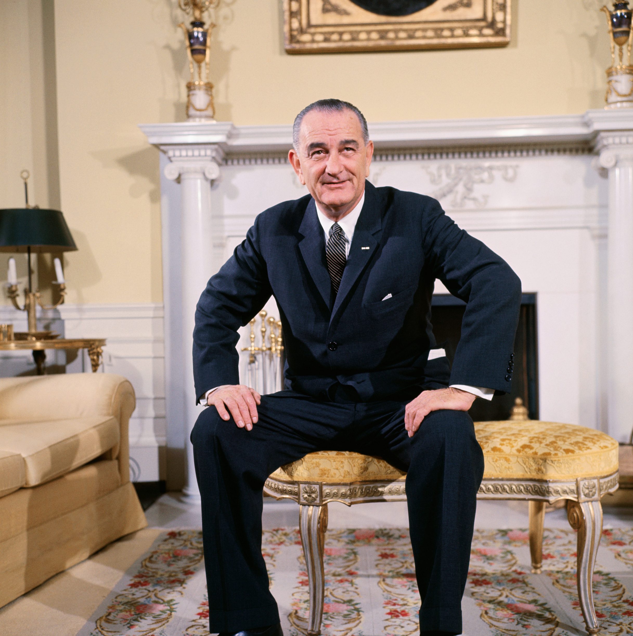 Lyndon B. Johnson sits on a bench in a formal room with ornate decor. He is wearing a dark formal suit