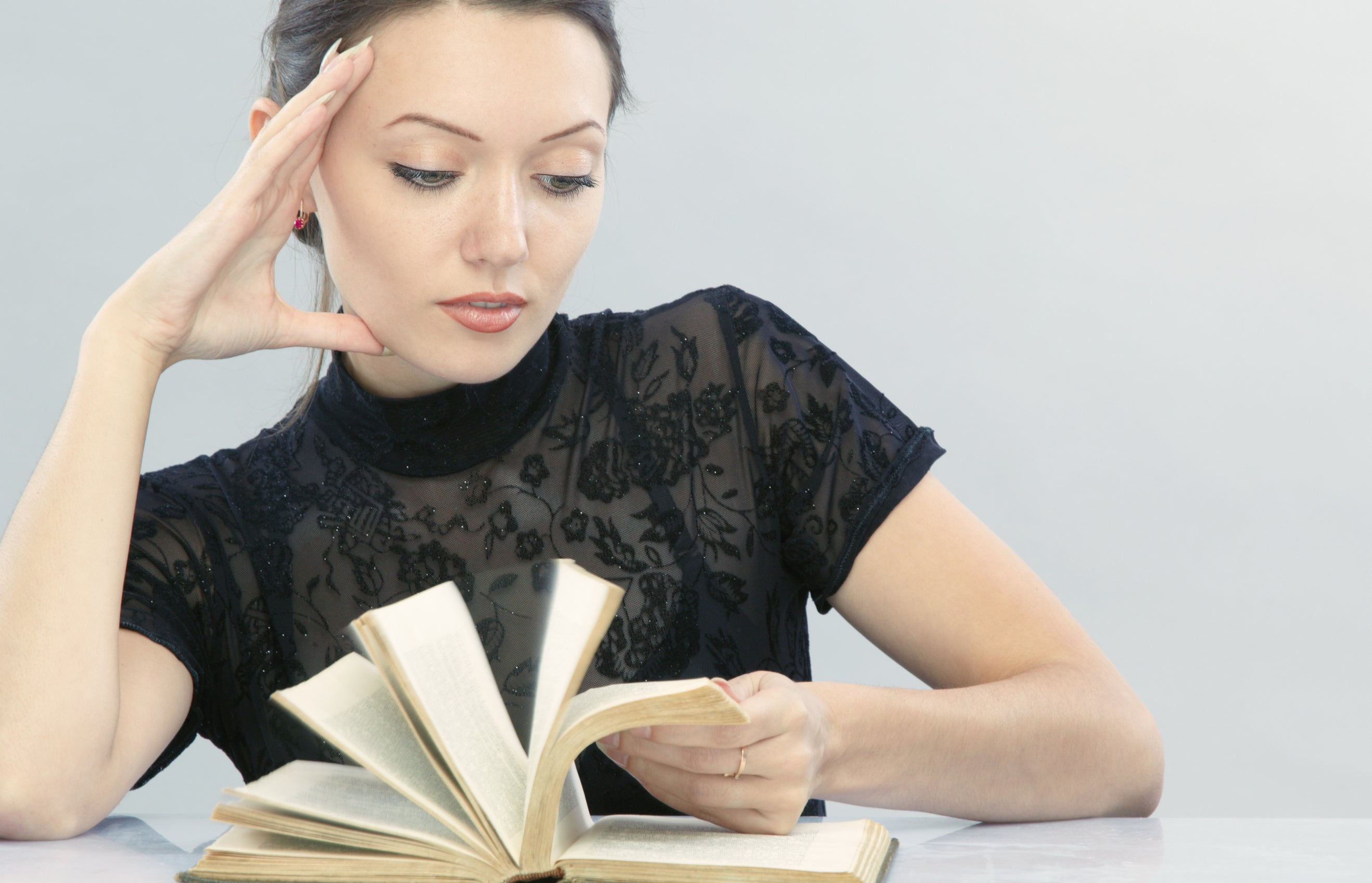 A woman in a lace top sits at a table, with one hand resting on her face and the other flipping through the pages of an open book
