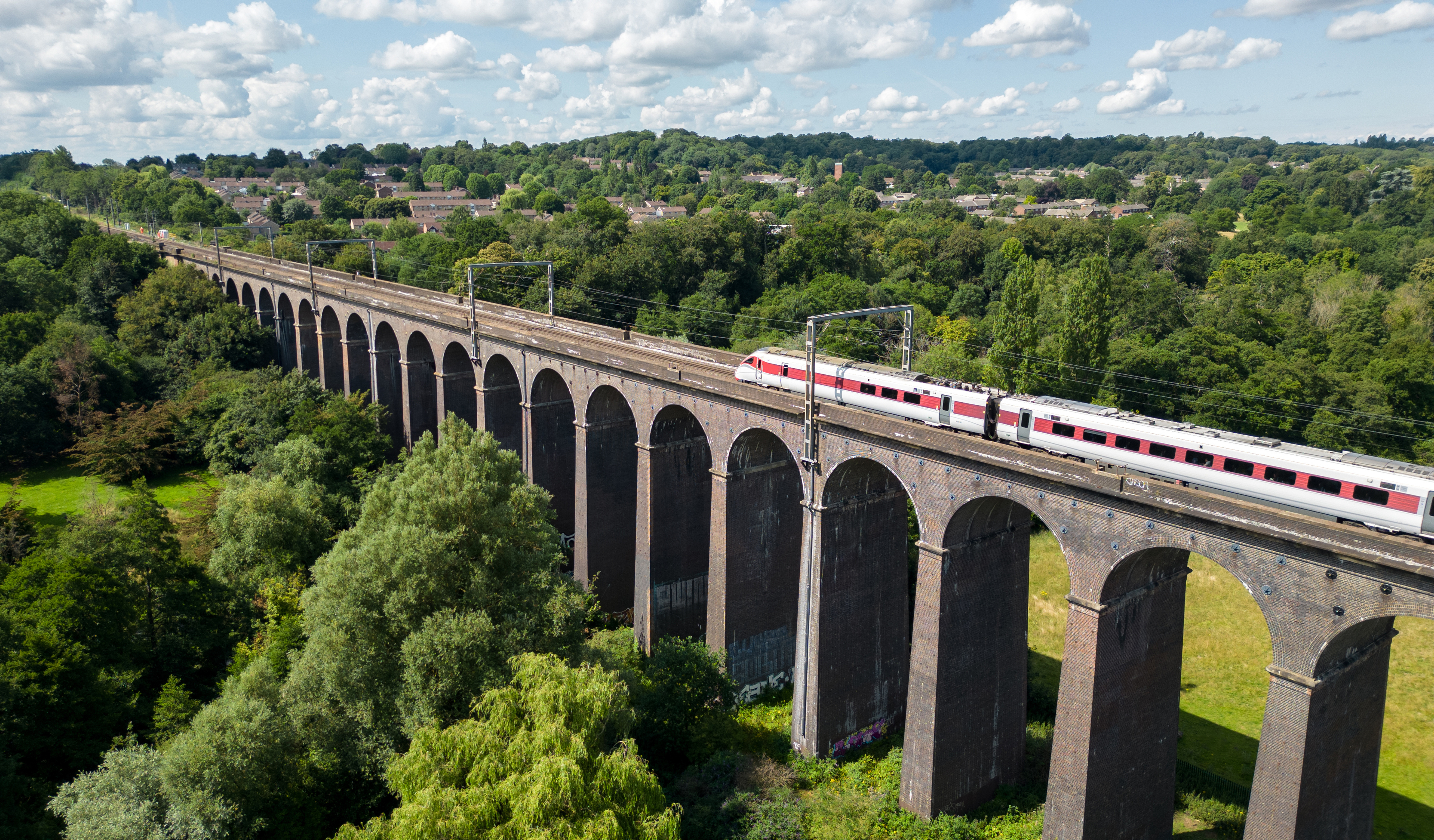 A train travels across a long brick viaduct surrounded by lush greenery and trees with a background of a small, distant town under a partly cloudy sky