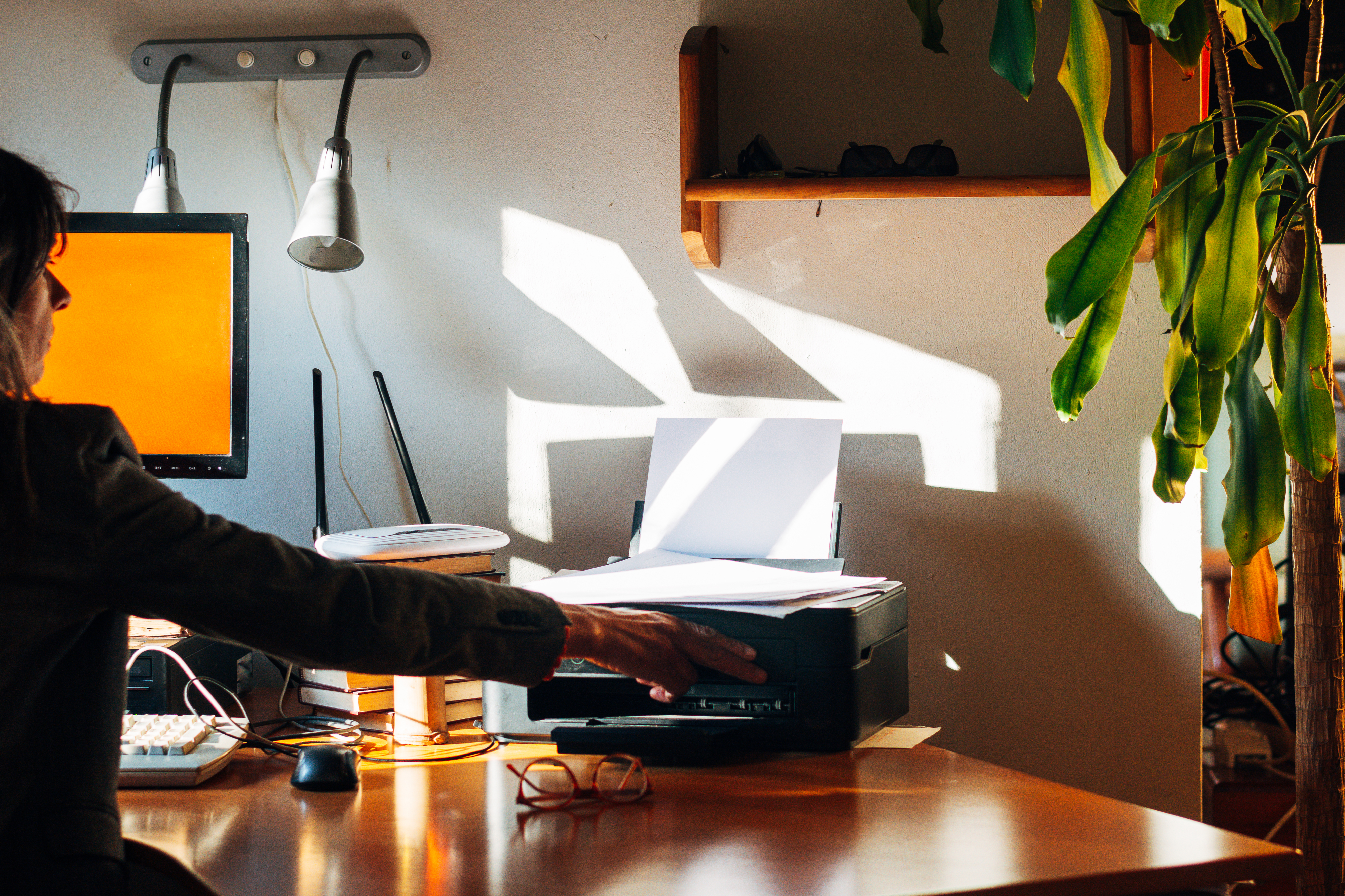 A person reaches for a printed paper from a printer on a cluttered desk with a computer, books, glasses, and a plant nearby