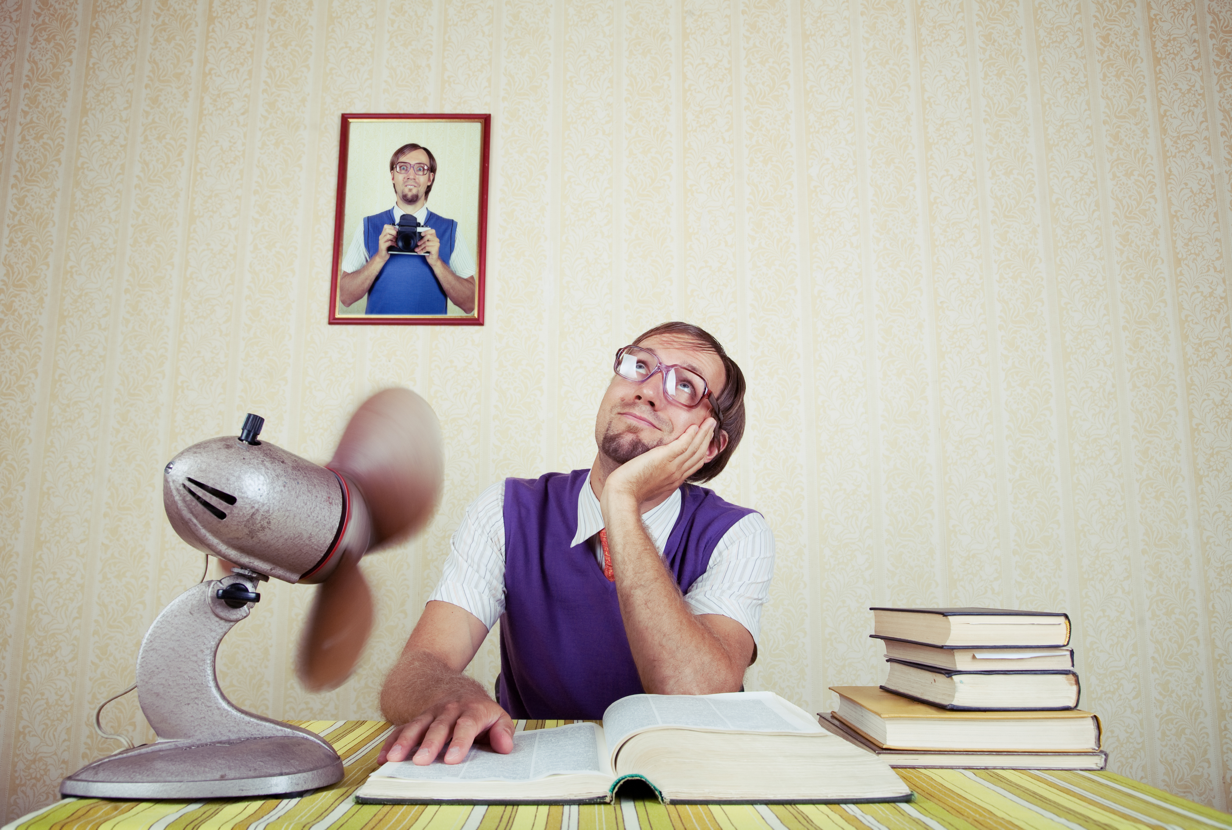 A man with glasses and vintage-style clothing sits at a desk with open books, looking pensive. A framed photo of him holding a camera is on the wall behind