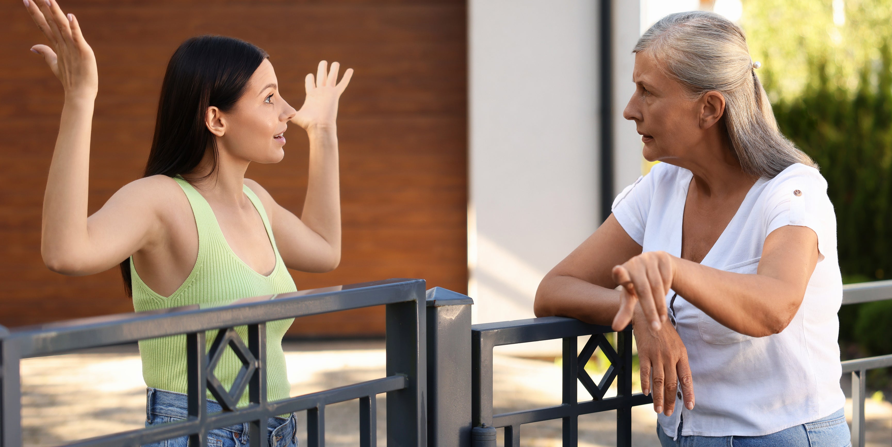 Two women, one younger with long dark hair in a sleeveless top and jeans, and one older with graying hair in a white top and jeans, talking over a metal fence