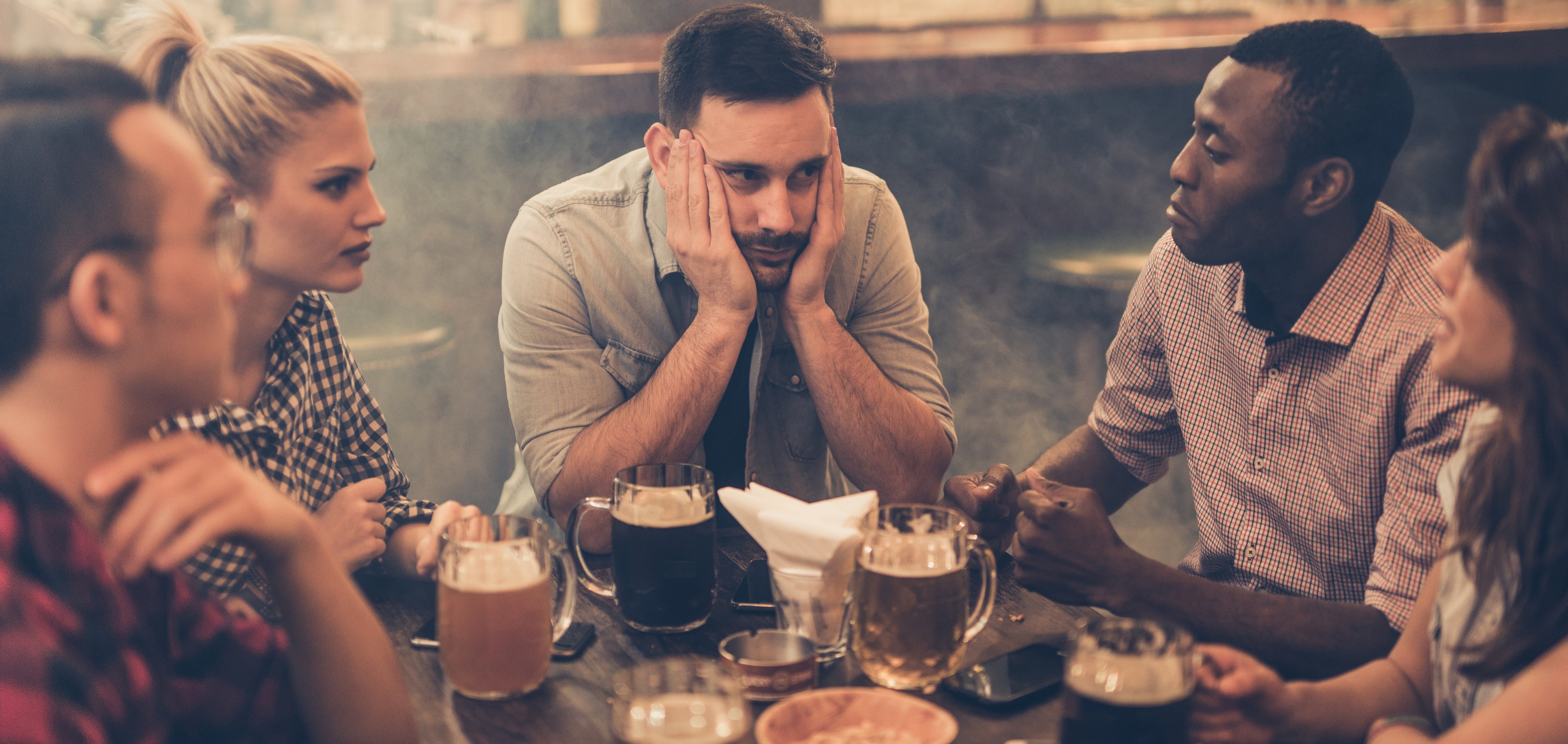 Five people sitting around a table with drinks in a bar. A man in the middle looks bored or stressed, resting his head on his hands.