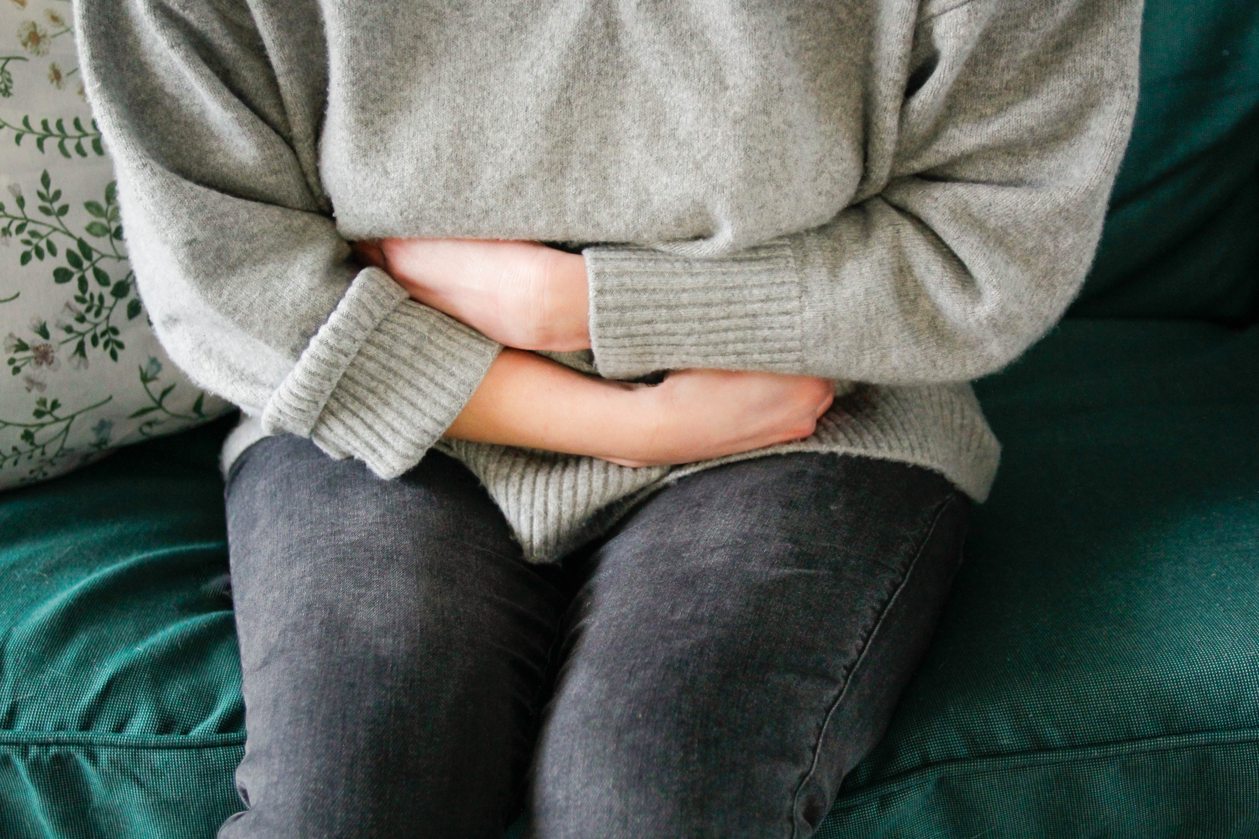 Person seated on a couch, wearing a sweater and jeans, with their arms crossed over their abdomen
