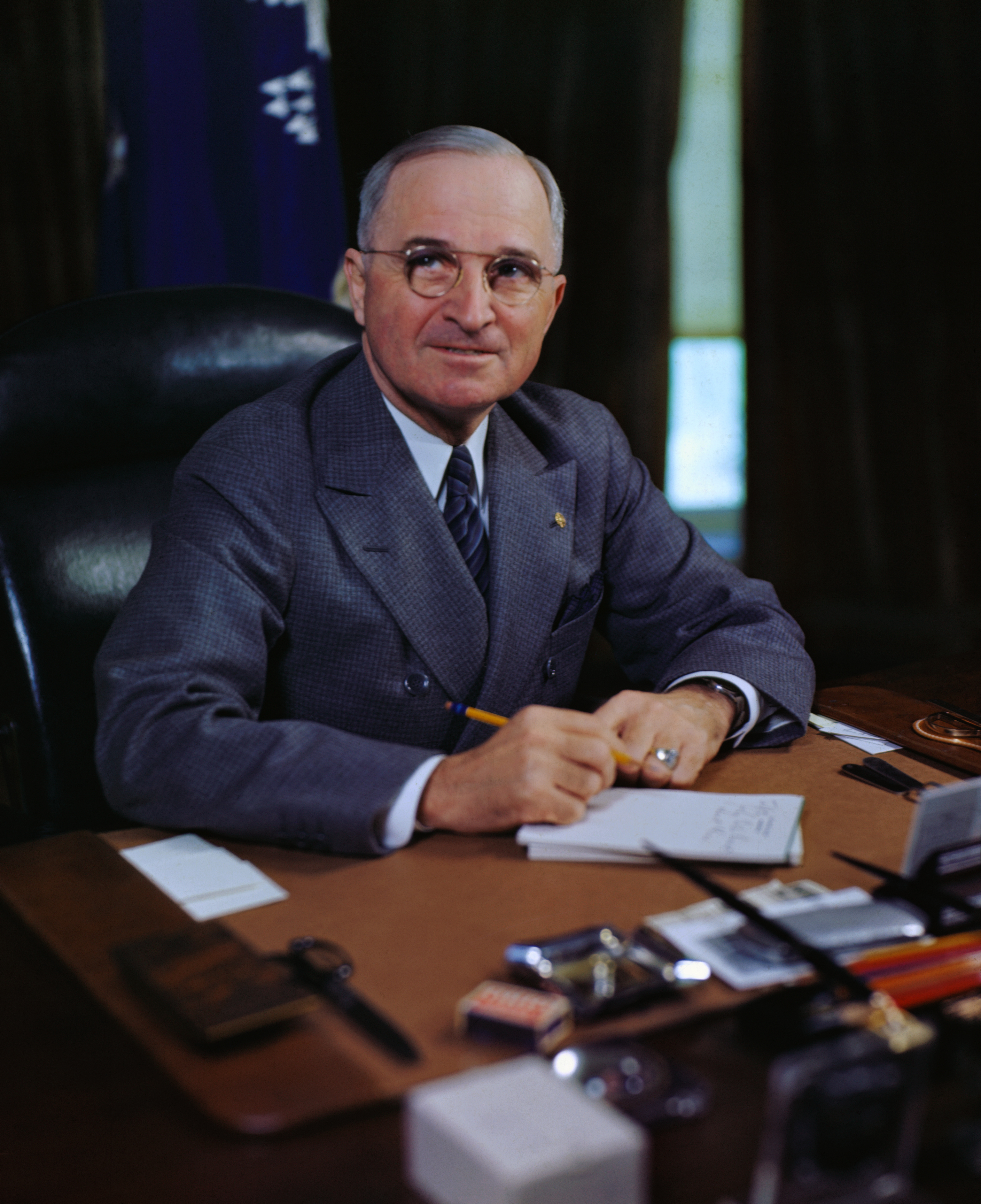 Harry S. Truman sits at his desk, wearing a formal suit and glasses, holding a pen and looking upward thoughtfully