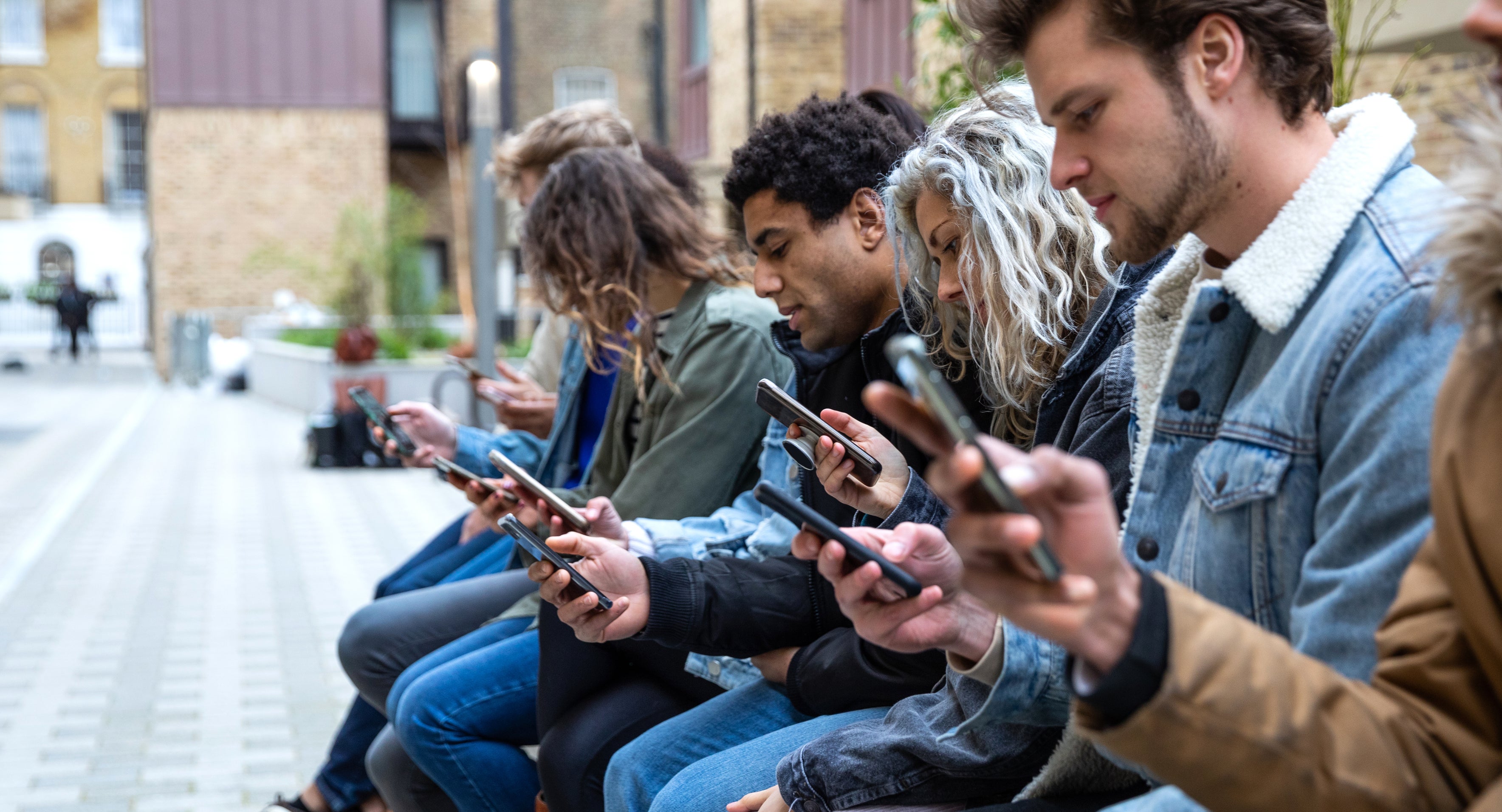 A diverse group of men and women sit on an outdoor bench, all focused on their smartphones