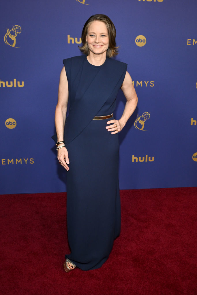 Jodie Foster at the Emmys, wearing a sleeveless, floor-length dress with minimalistic design. She’s smiling and posing on the red carpet