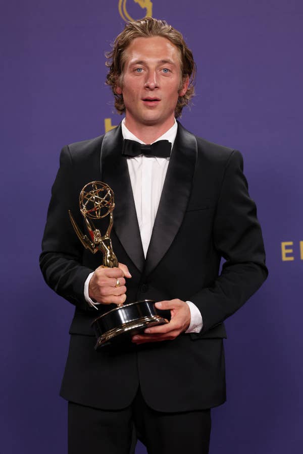 Man in a black tuxedo holding an Emmy Award against a purple backdrop. Names of persons unknown