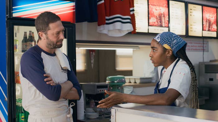 Man and woman in a diner, woman speaking with hand gesture. Menu boards in the background
