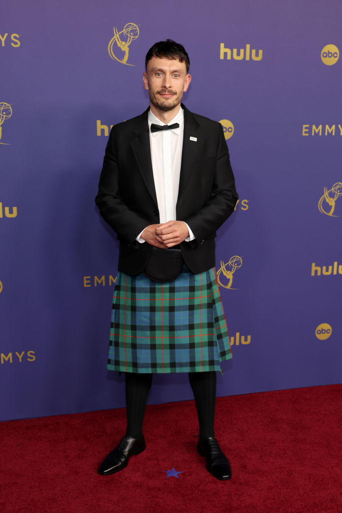 Person wearing a tuxedo jacket with bow tie and a plaid kilt on a red carpet at the Emmys event. Banners for Hulu and ABC are in the background