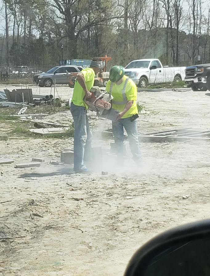 Two construction workers wearing neon safety vests and helmets operate a concrete saw on a dusty worksite with vehicles and trees in the background