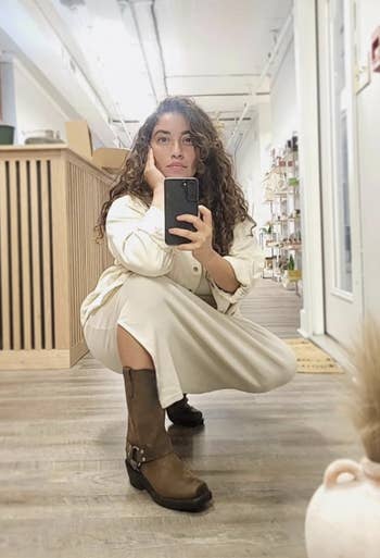 A pistillate successful a achromatic outfit and brownish boots takes a reflector selfie portion squatting successful a country with shelves and a counter