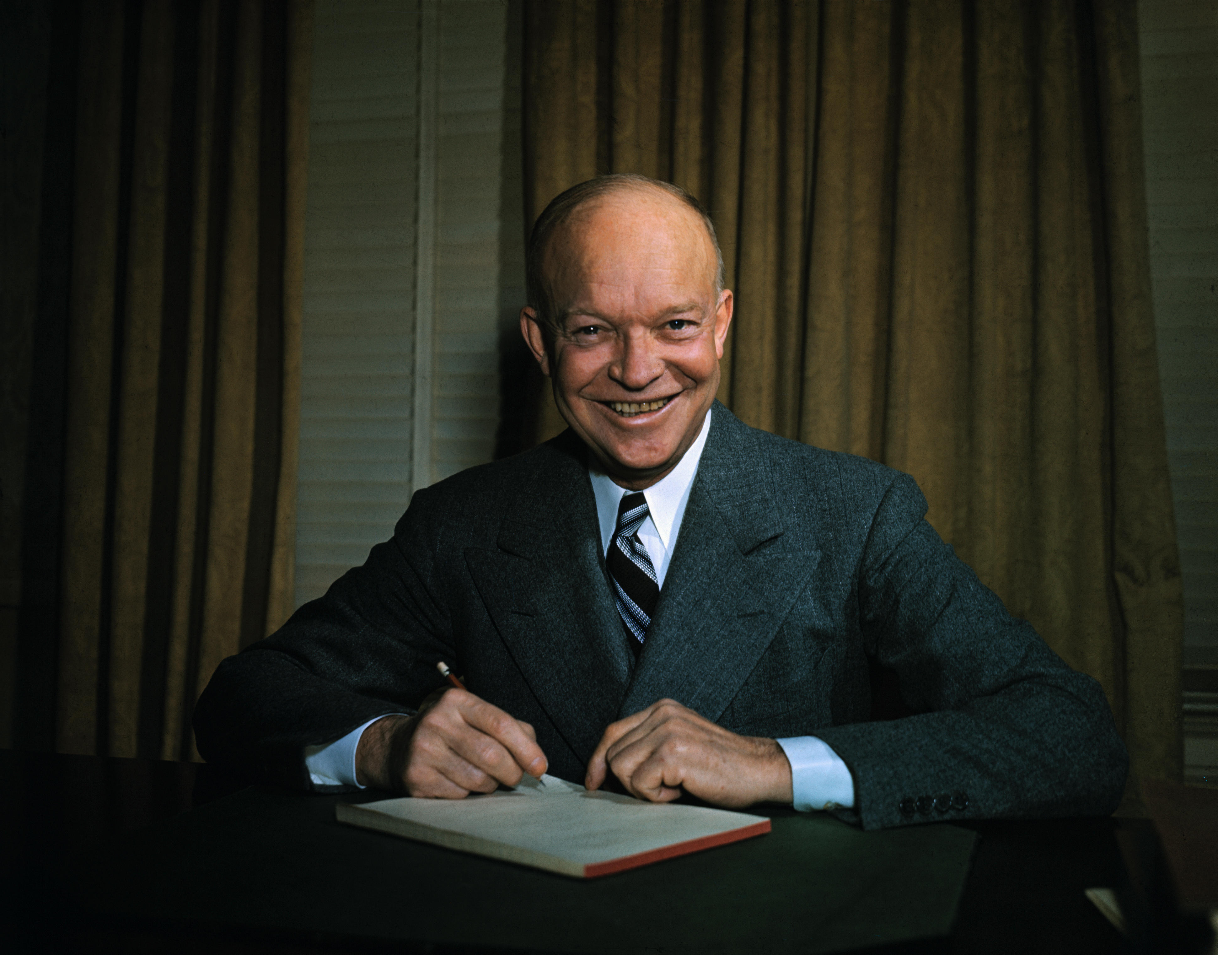 Dwight D. Eisenhower sits at a desk, smiling, in a suit with a tie. He is writing on a document