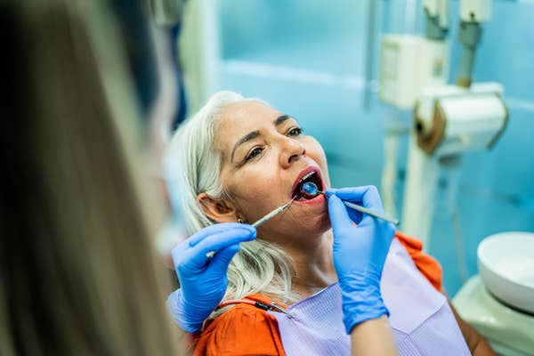 Kobieta jest u dentysty, przechodzi przegląd stomatologiczny. Dentysta w niebieskich rękawiczkach bada jej jamę ustną narzędziami dentystycznymi.
