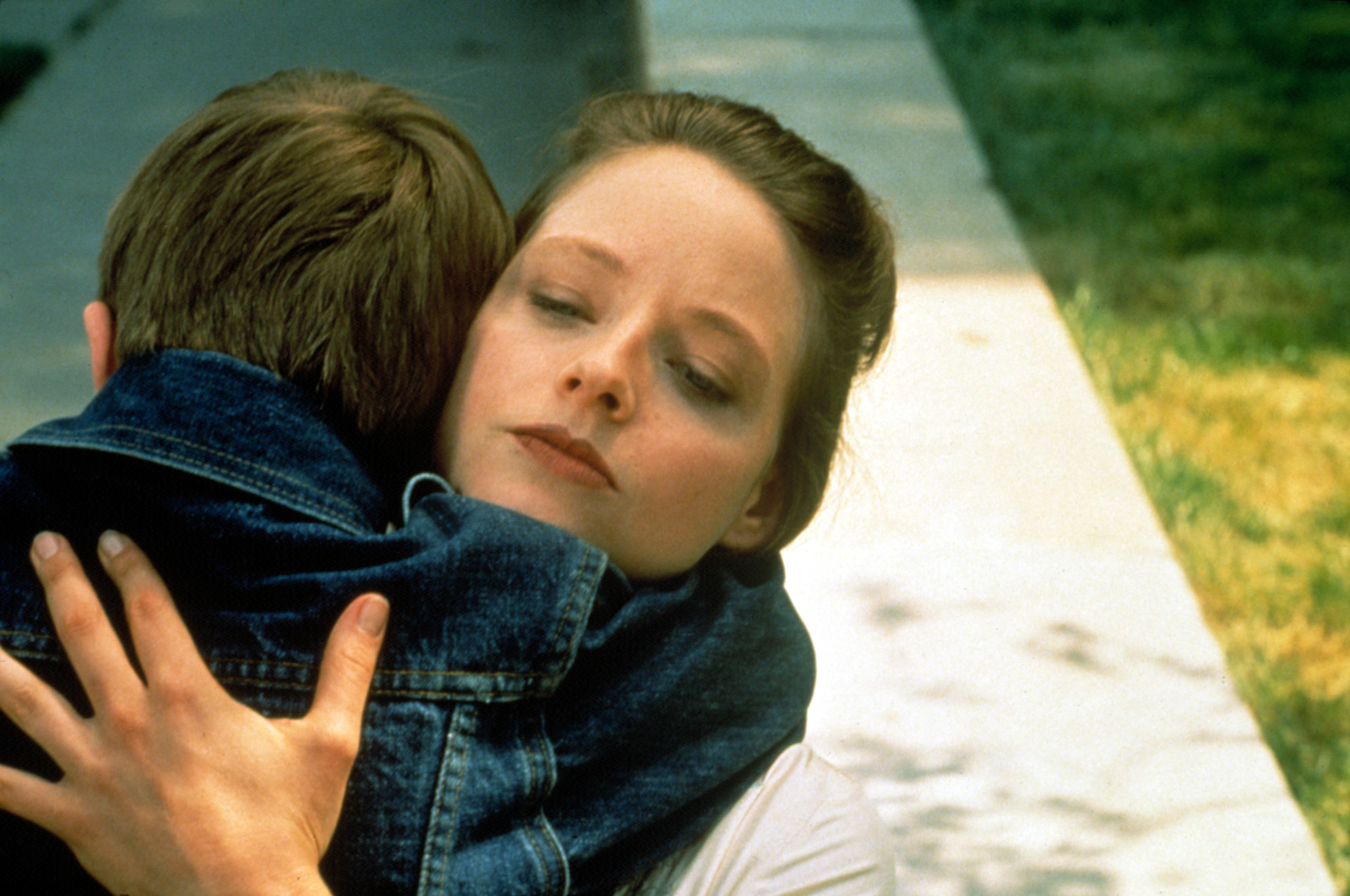 Jodie Foster embraces a young child, both wearing denim jackets, in an outdoor setting