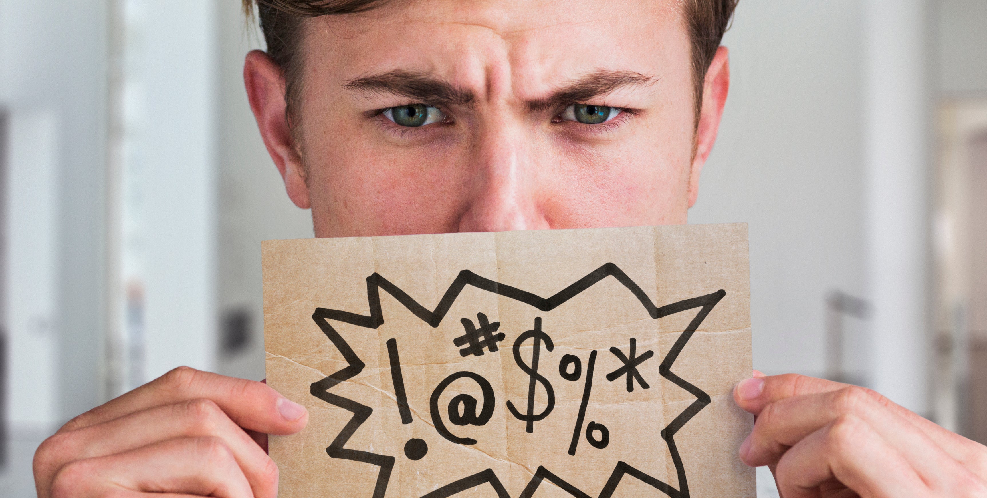 A person holds up a sign with various expletive symbols (!, @, #, $, %, *) in frustration, covering their mouth