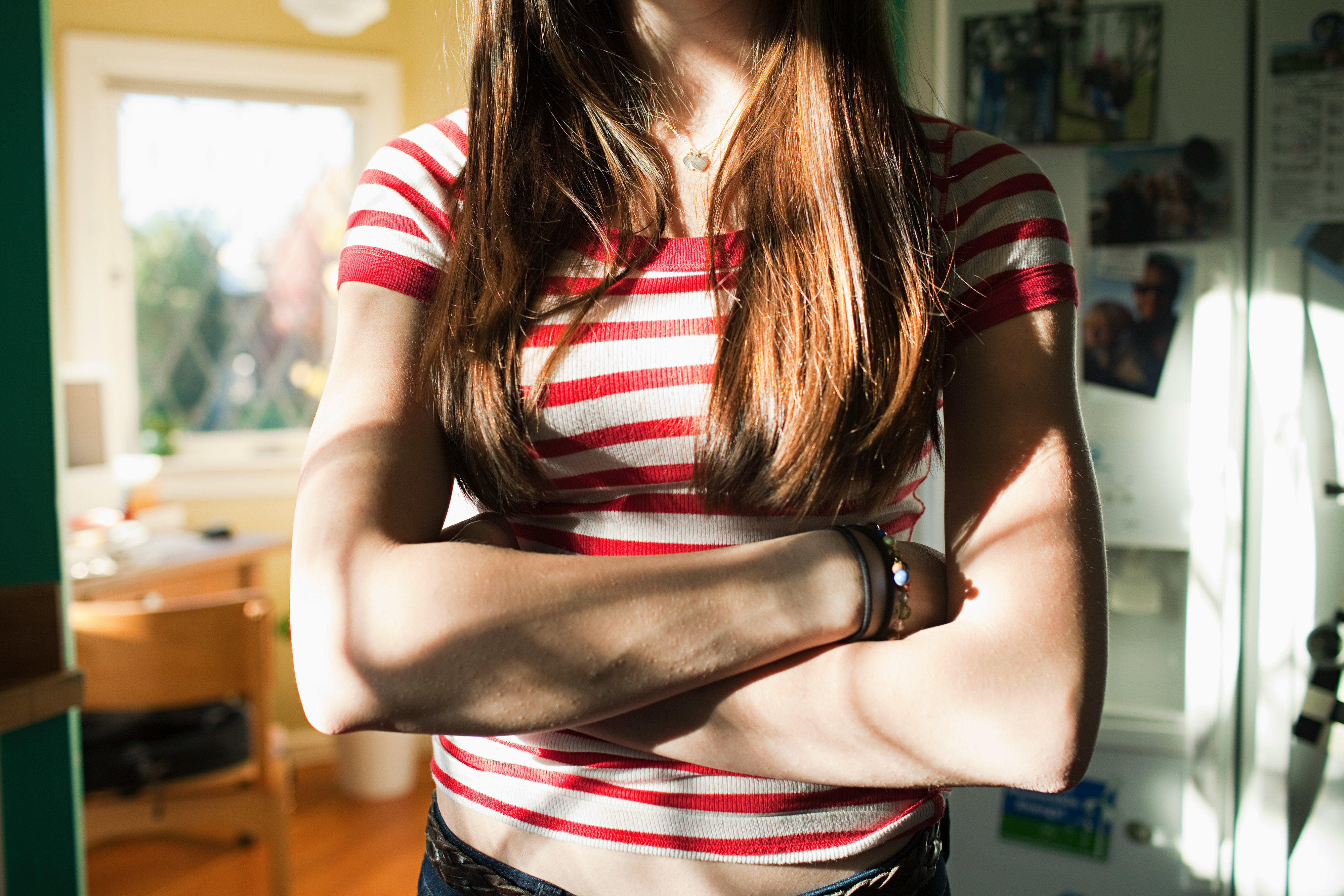A person with long hair is standing indoors with arms crossed, wearing a short-sleeved, red and white striped shirt. No face is visible