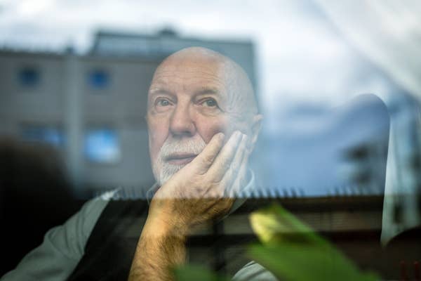 Starszy mężczyzna patrzy zamyślony przez okno, opierając dłoń na policzku. W tle widać rozmazany budynek i trochę zieleni.