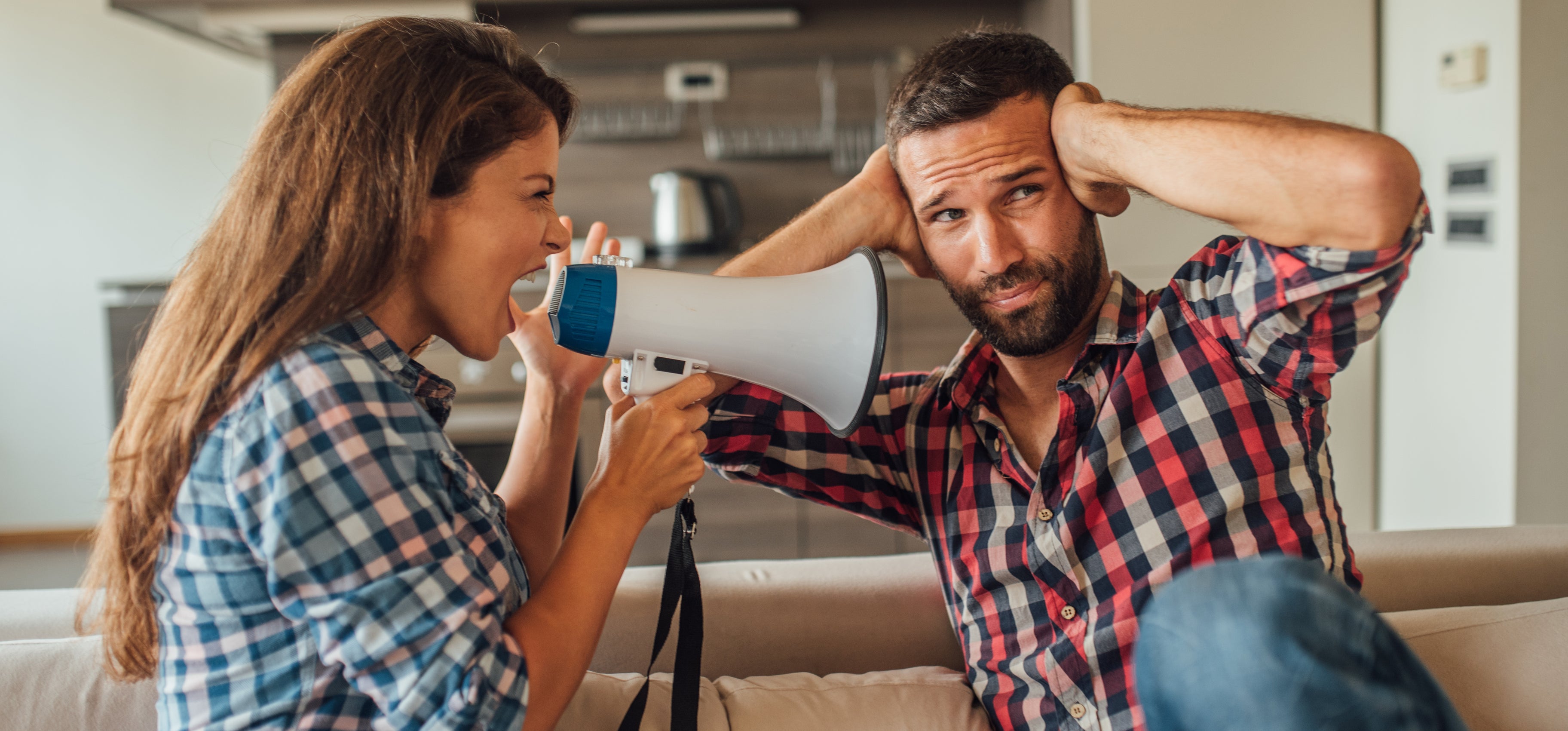 A woman uses a megaphone to shout at a man who is covering his ears. Both are casually dressed in plaid shirts. The setting appears to be a kitchen