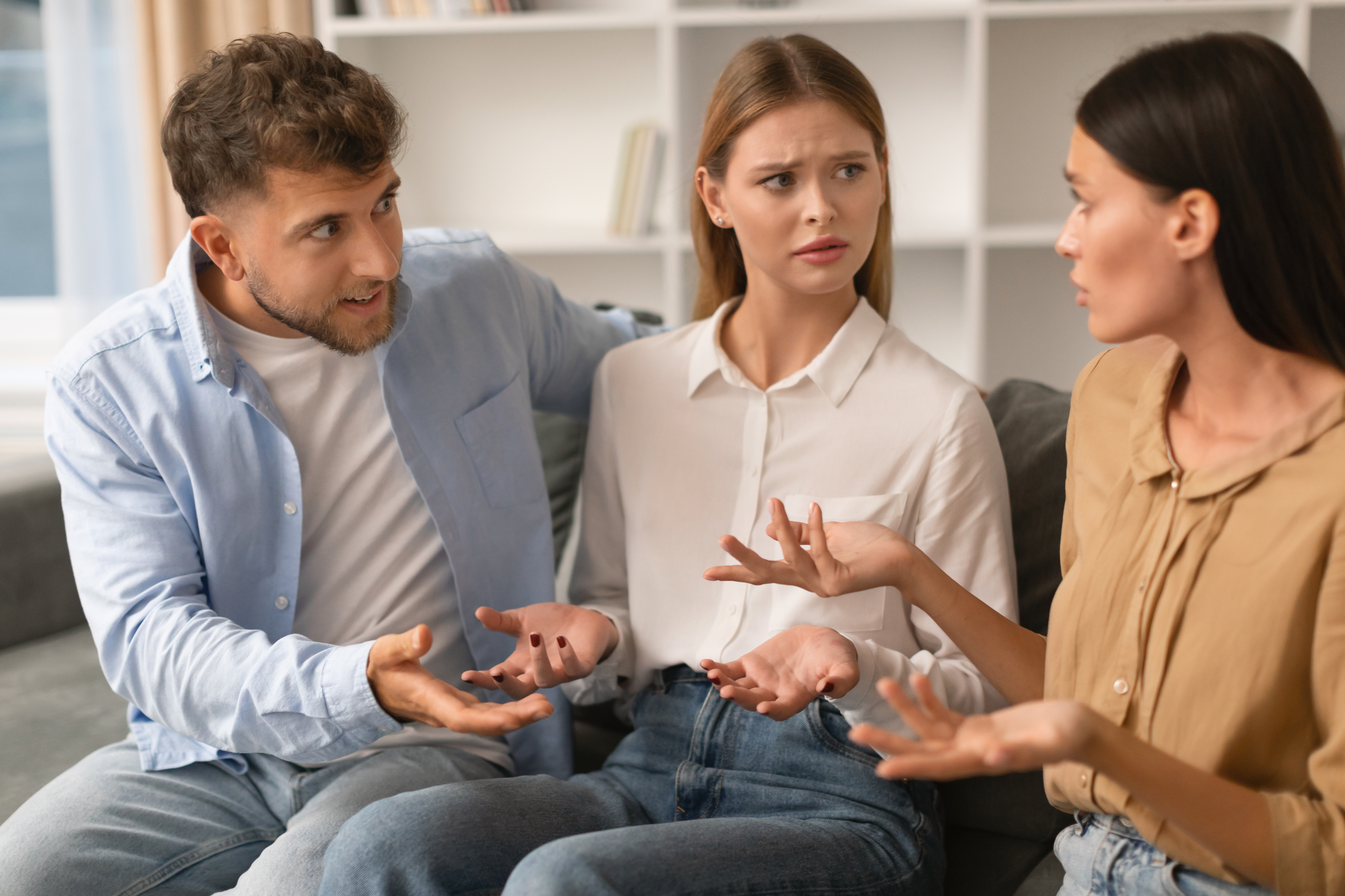 Three people are sitting indoors having a serious conversation. The woman in the middle looks concerned, while the man and another woman are gesturing animatedly