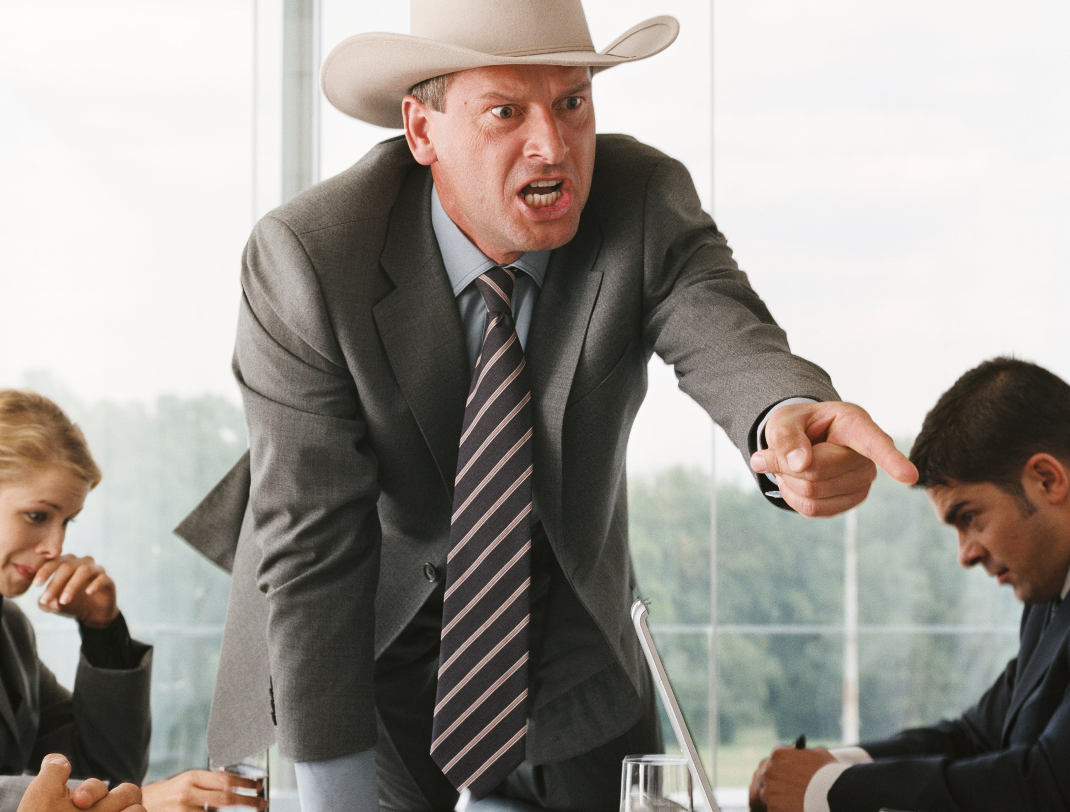 Man in a suit and cowboy hat stands on a conference table, angrily pointing. Four people in suits sit around the table looking shocked