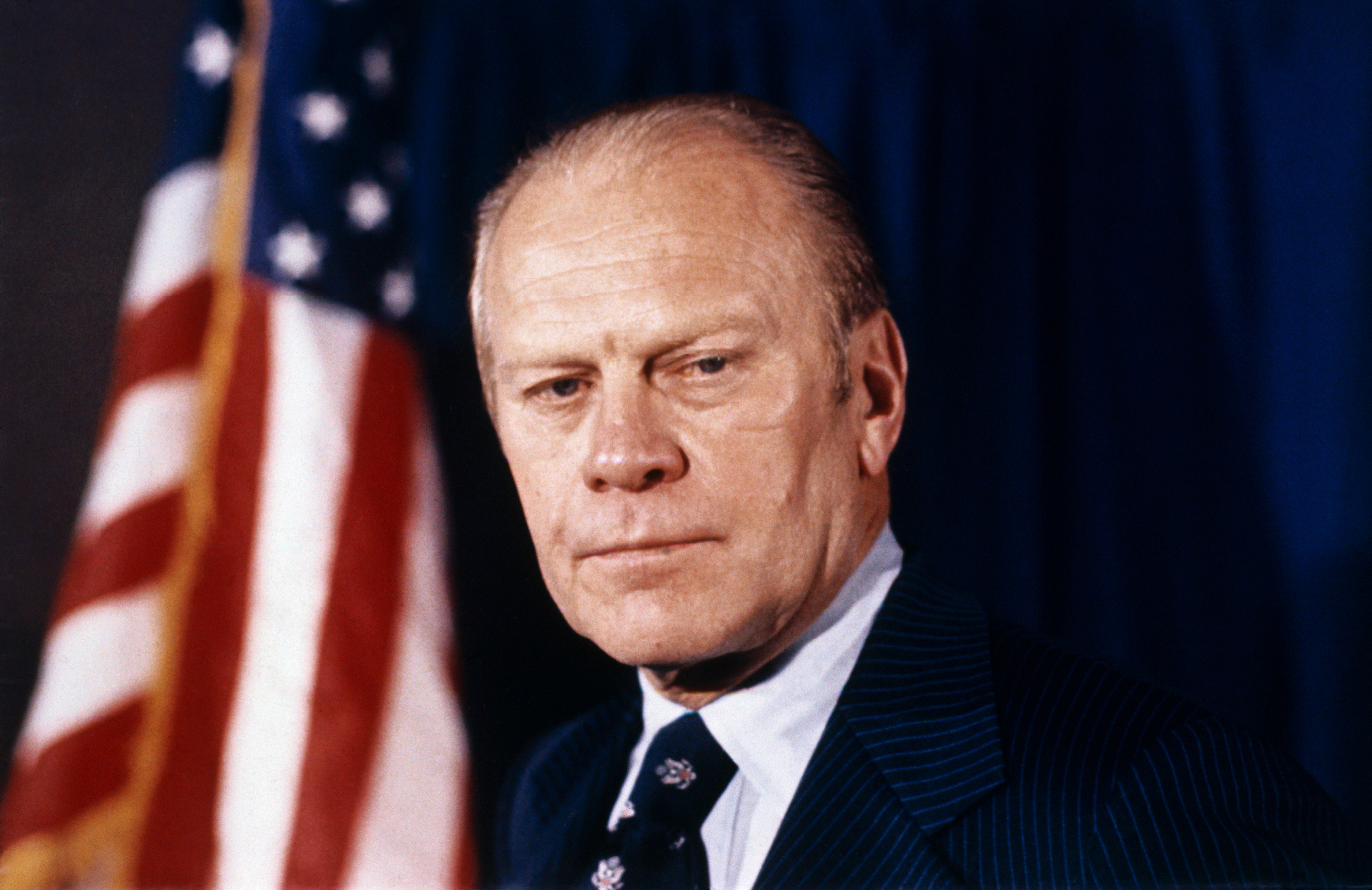 Gerald Ford stands next to an American flag, wearing a suit with a pinstriped tie