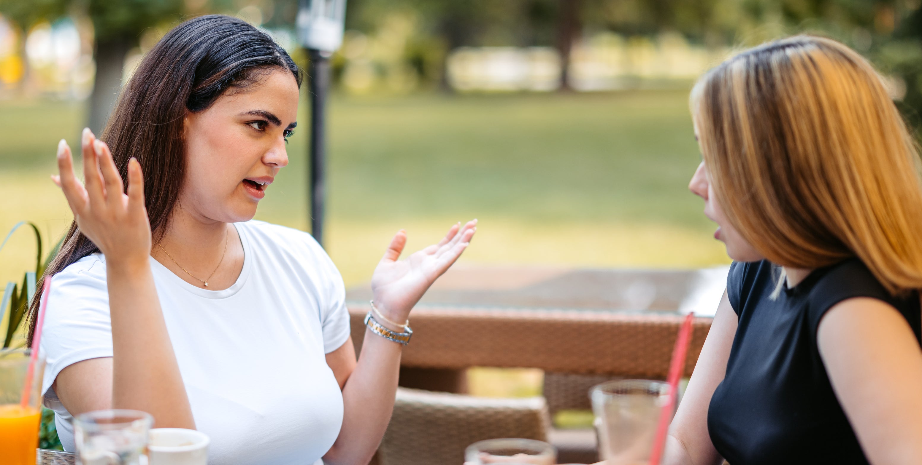 Two women sit at an outdoor cafe table, engaged in a lively conversation with drinks and food in front of them
