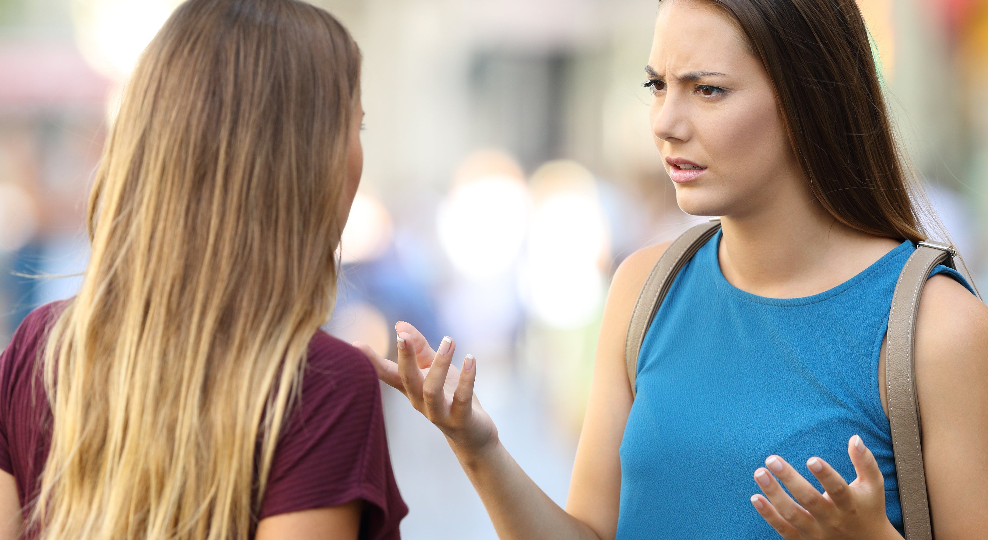 Two women having a serious conversation outdoors. Both appear animated; one is wearing a blue top, the other a maroon top. Their expressions seem intense