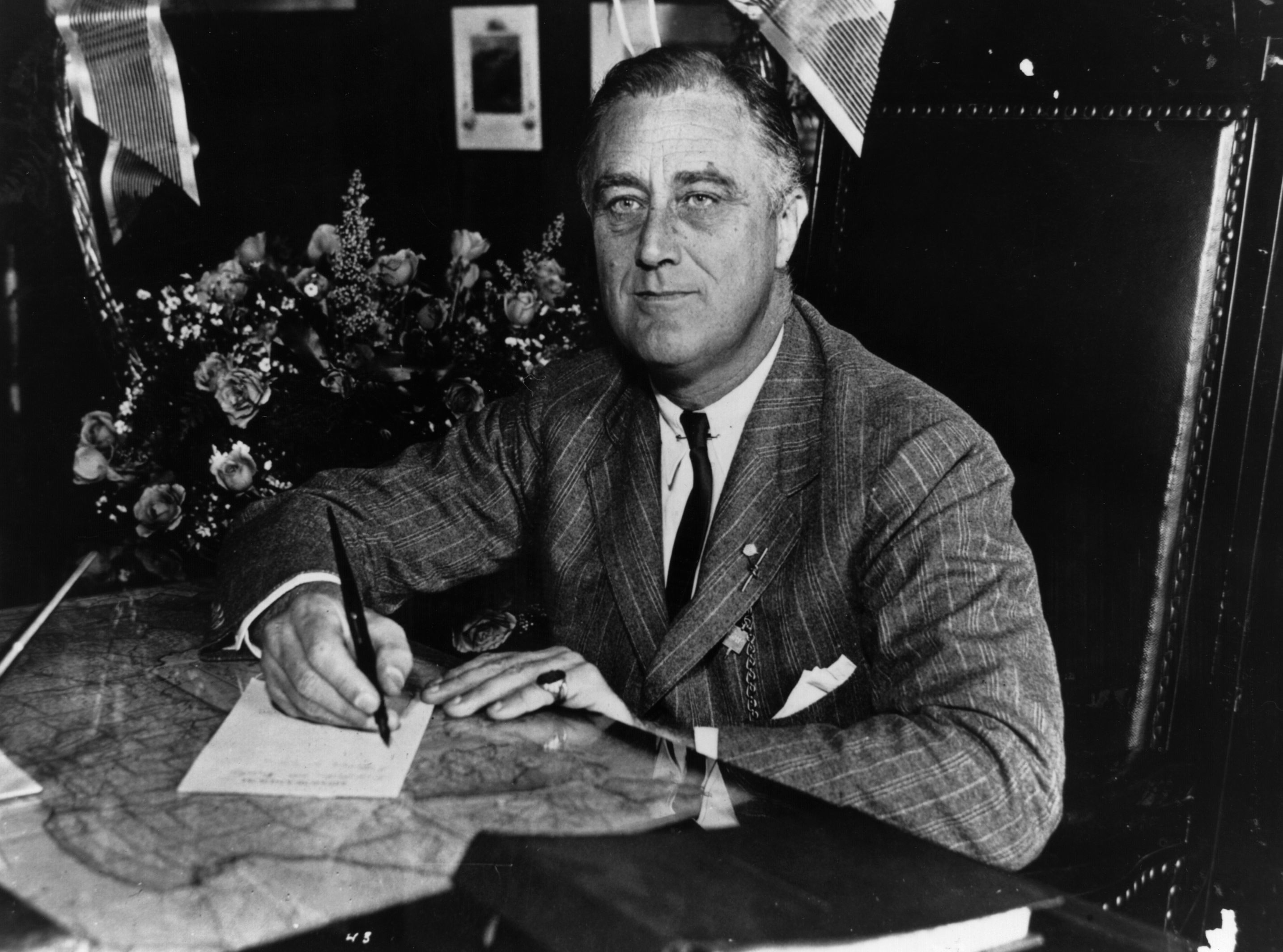 Franklin D. Roosevelt sits at a desk holding a pen, with flowers and papers around him