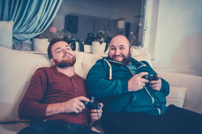 Two men relax on a couch playing video games, one with a beard and red shirt, the other with a bald head and blue hoodie, smiling and sharing a moment