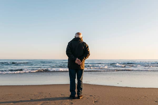 Osoba stoi samotnie na plaży, patrzy na ocean, trzymając ręce splecione z tyłu.