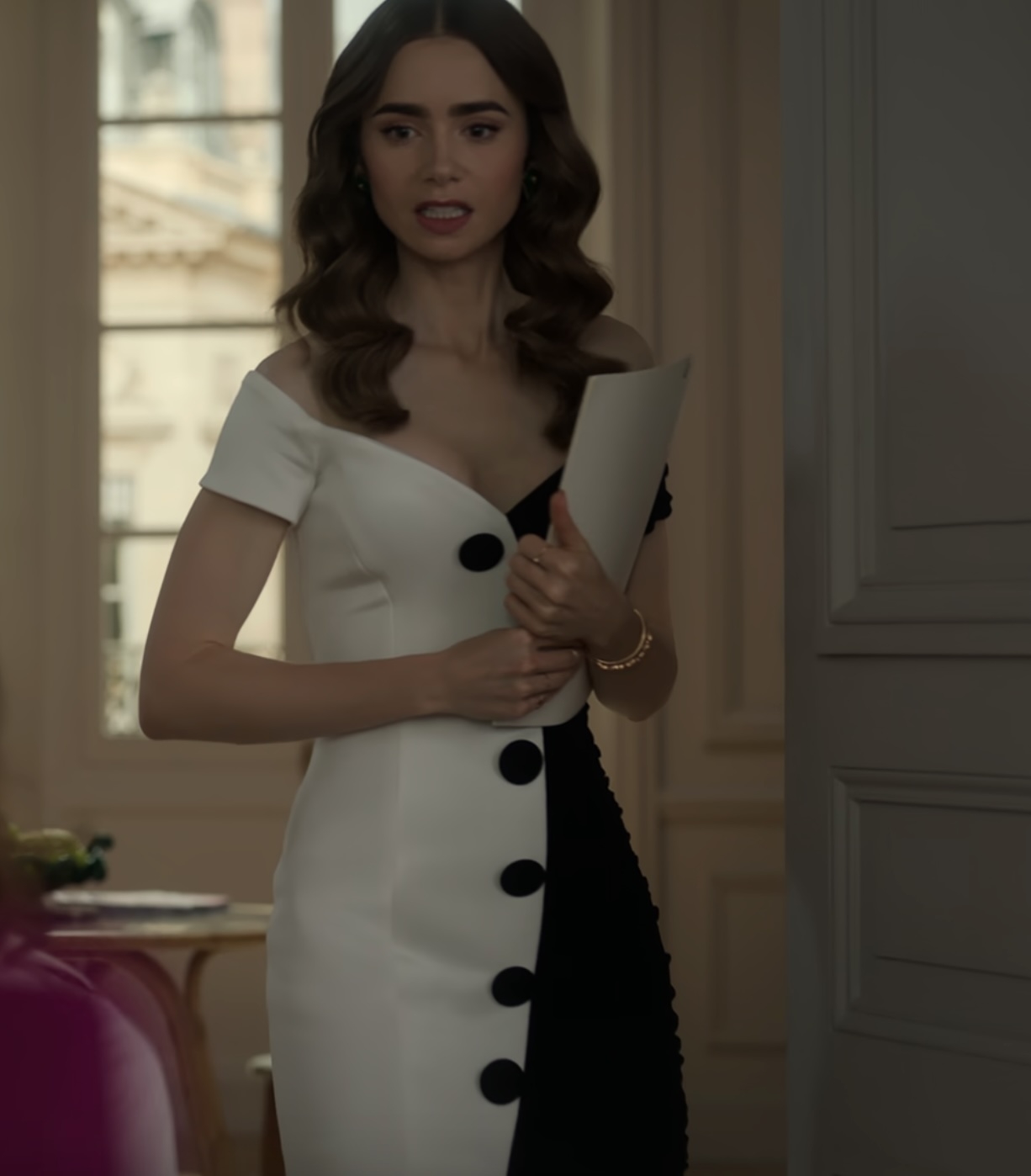 Lily Collins dressed in a black and white, off-shoulder dress with large round buttons, holds a folder and looks ahead, standing in an indoor setting