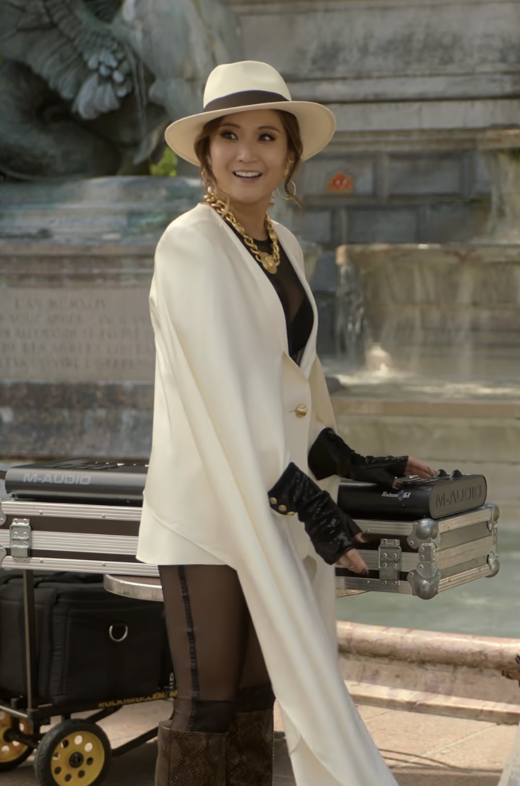 Ashley Park at an event, wearing a stylish white cape, black gloves, patterned leggings, gold accessories, and a hat, while standing beside equipment cases