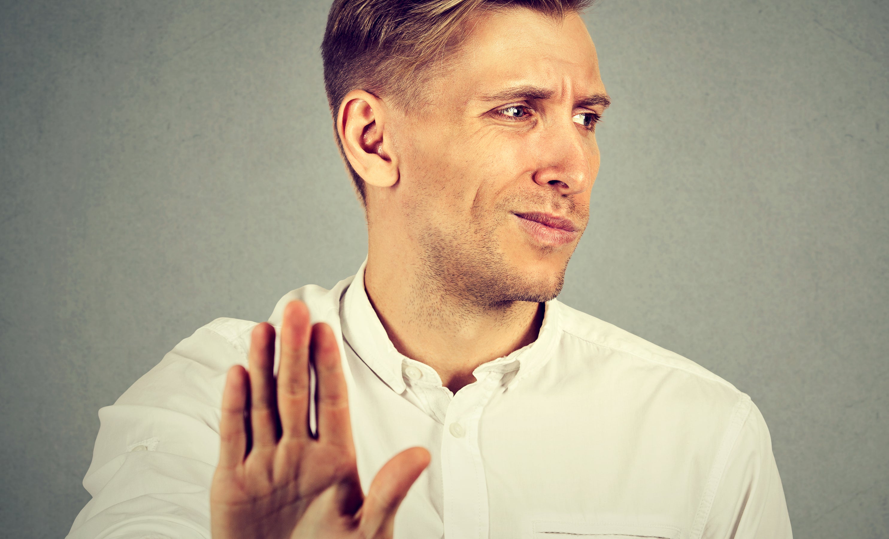 A man in a white shirt stands against a plain background, holding his hand up with a serious expression