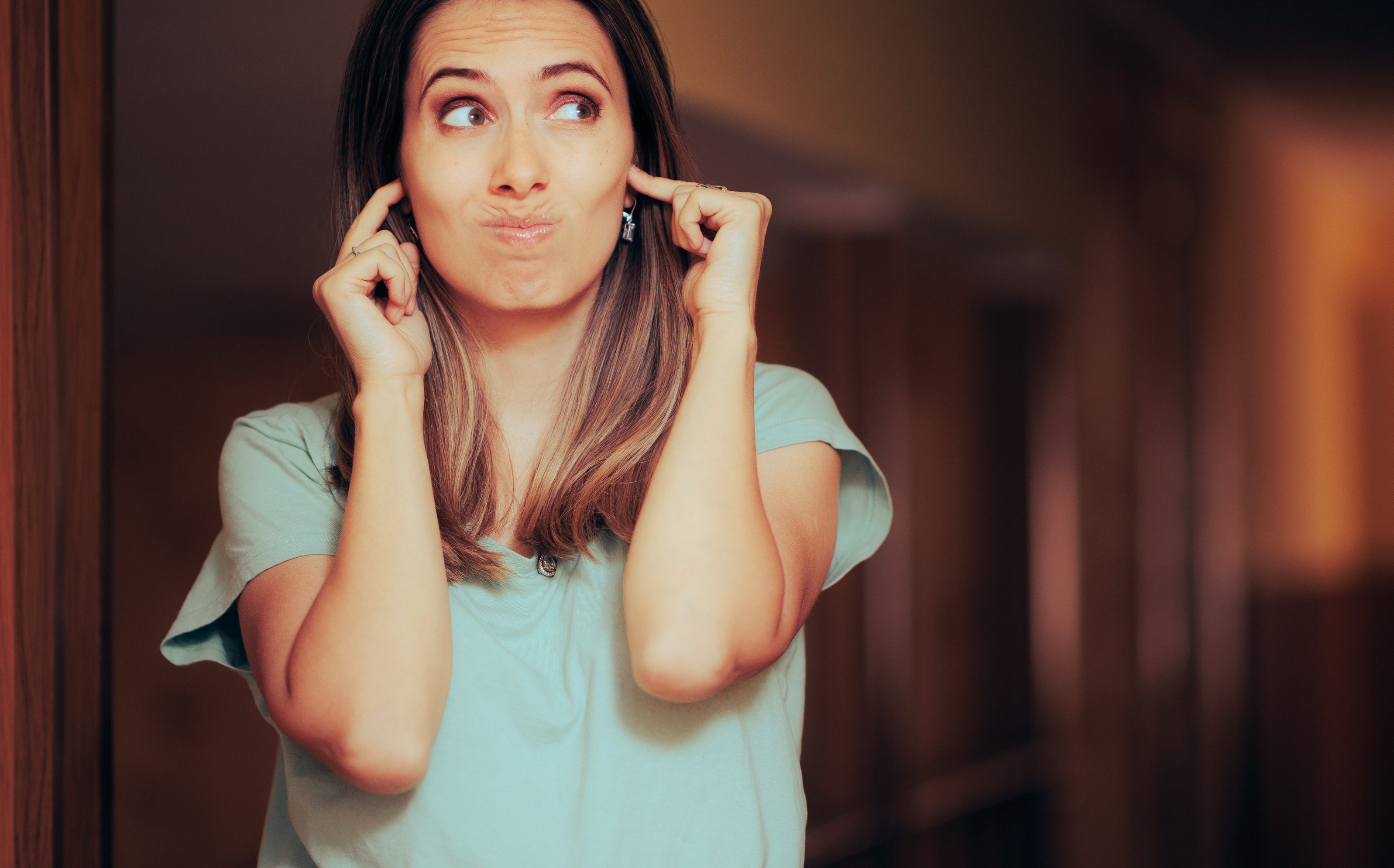 A woman with medium-length hair, wearing a casual top, stands in a hallway and covers her ears with her fingers while making a thoughtful expression