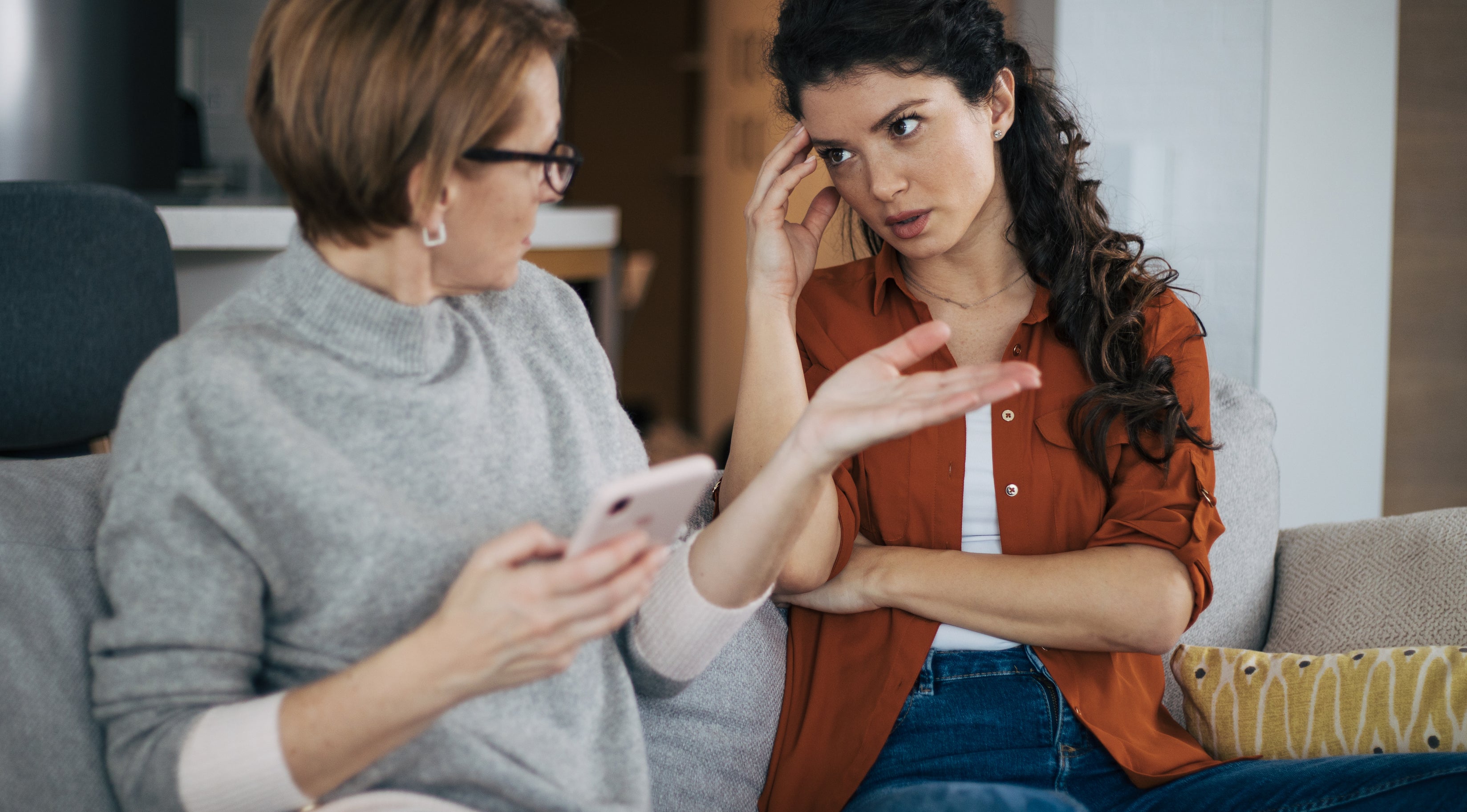 Two women, one older and one younger, sit on a couch engaged in a serious conversation. The older woman holds a phone; the younger woman appears concerned