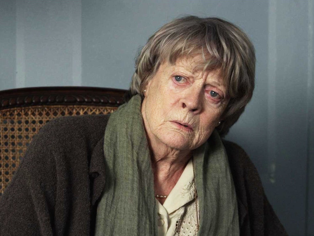 Maggie Smith sits on a wooden chair, looking pensive. She wears a loose coat and a scarf