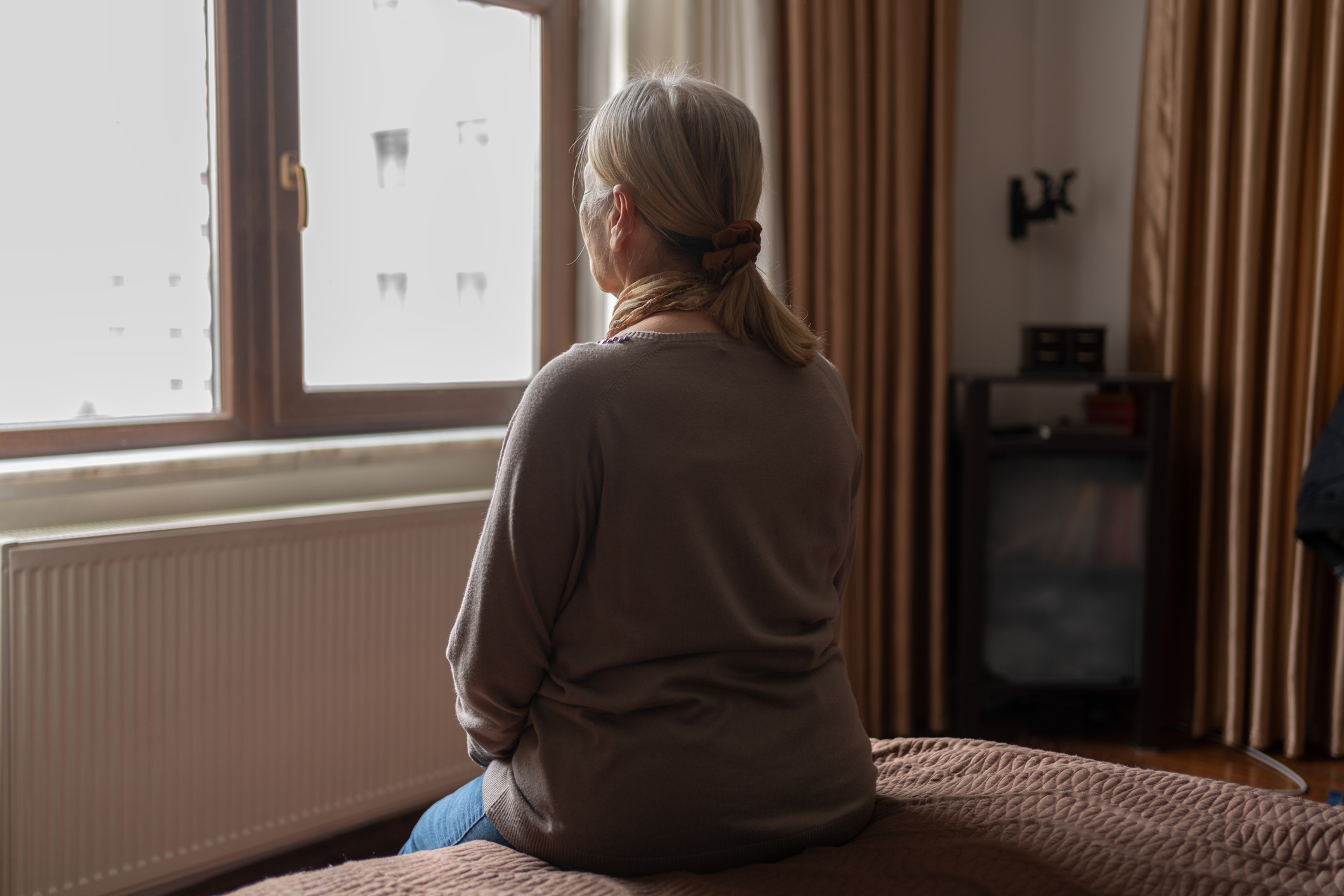 A woman with gray hair tied in a low ponytail sits on a bed, gazing out of a window