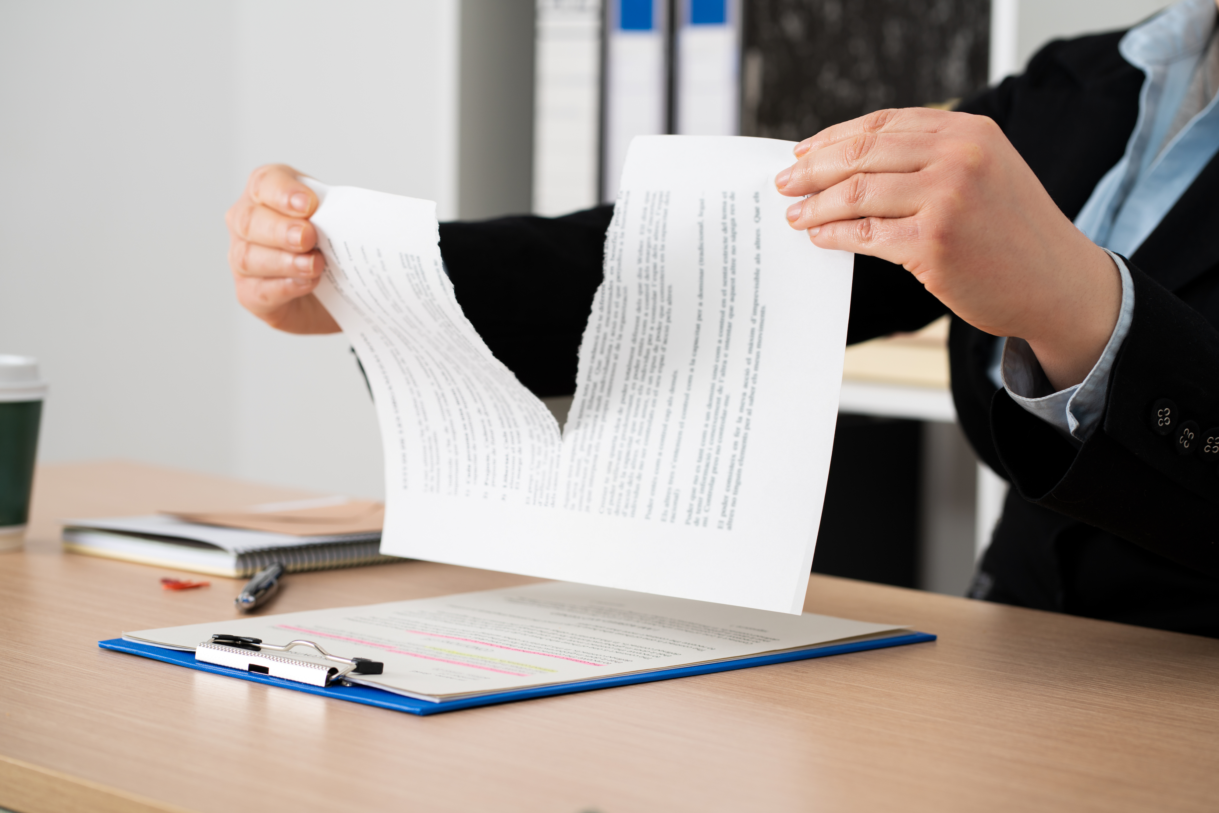 A person in business attire tears a document in half at a desk, with a clipboard and notebooks visible