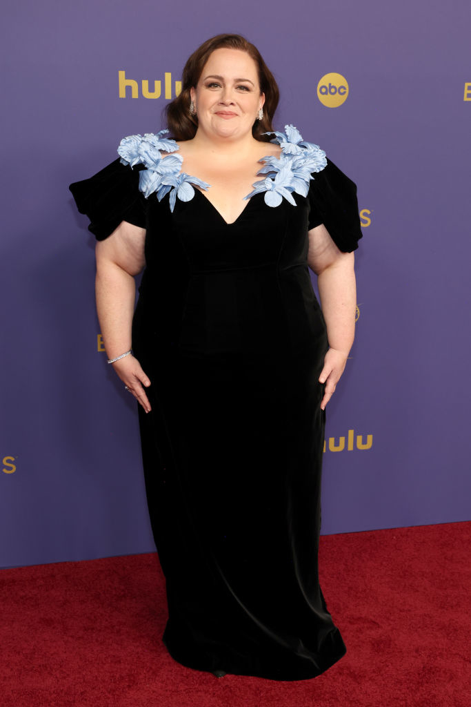 Nicole Byer on the red carpet wearing a black dress with blue floral details at the shoulders during a Hulu event